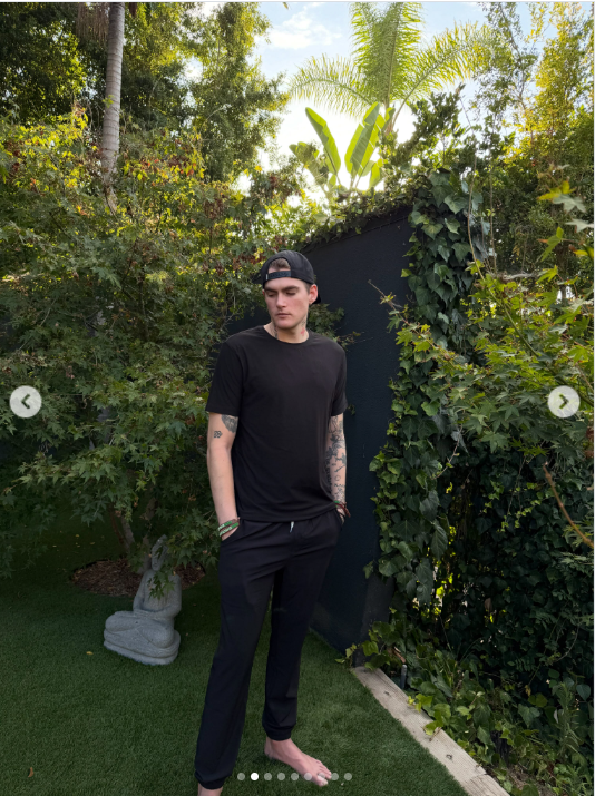 Presley Gerber se gira ligeramente hacia la cámara con una sonrisa discreta, de pie descalzo en el jardín, con los tatuajes de los brazos a la vista y una mano metida en el bolsillo. | Fuente: Instagram/presleygerber