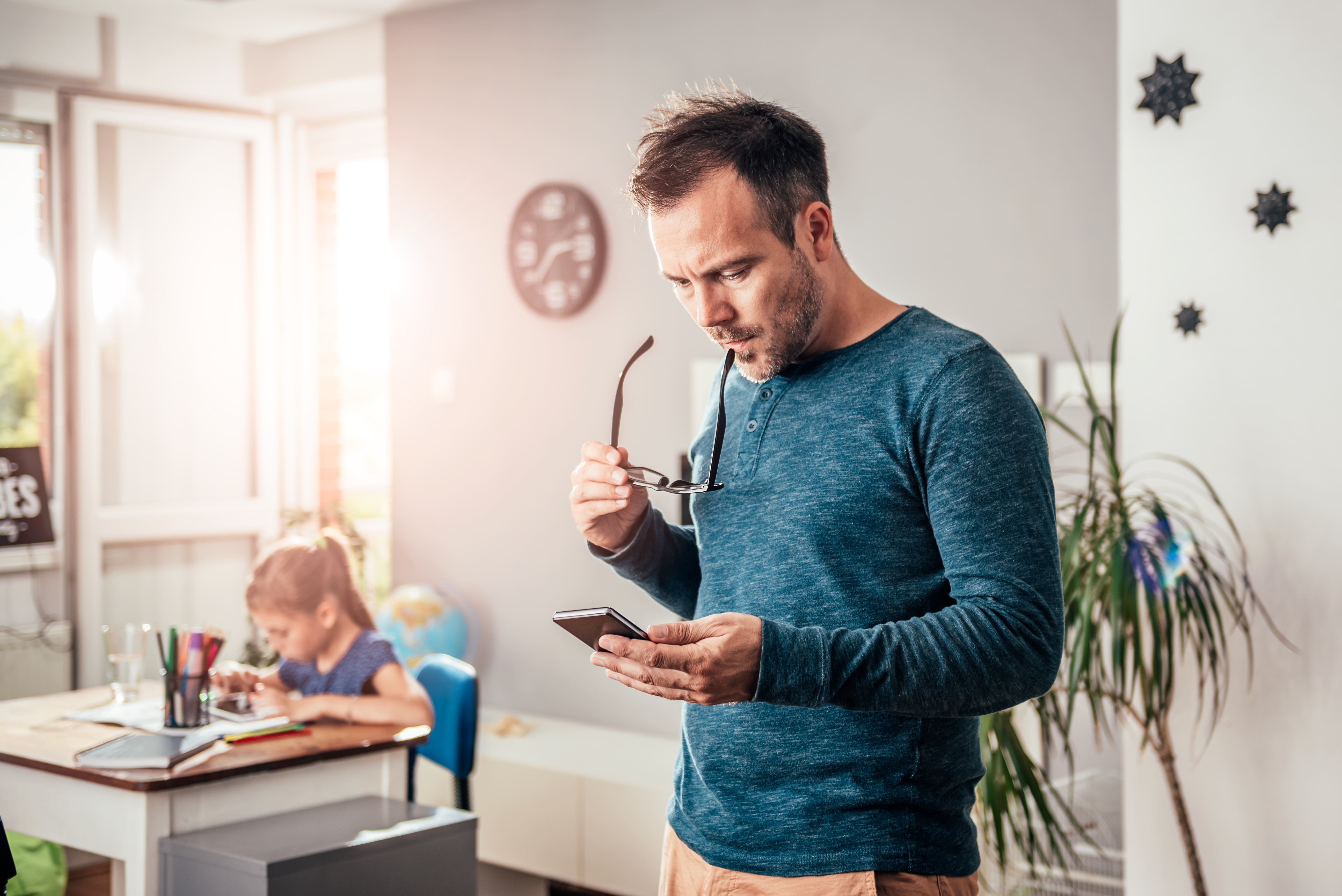 Hombre mirando su teléfono mientras una niña estudia en un escritorio | Foto: Shutterstock