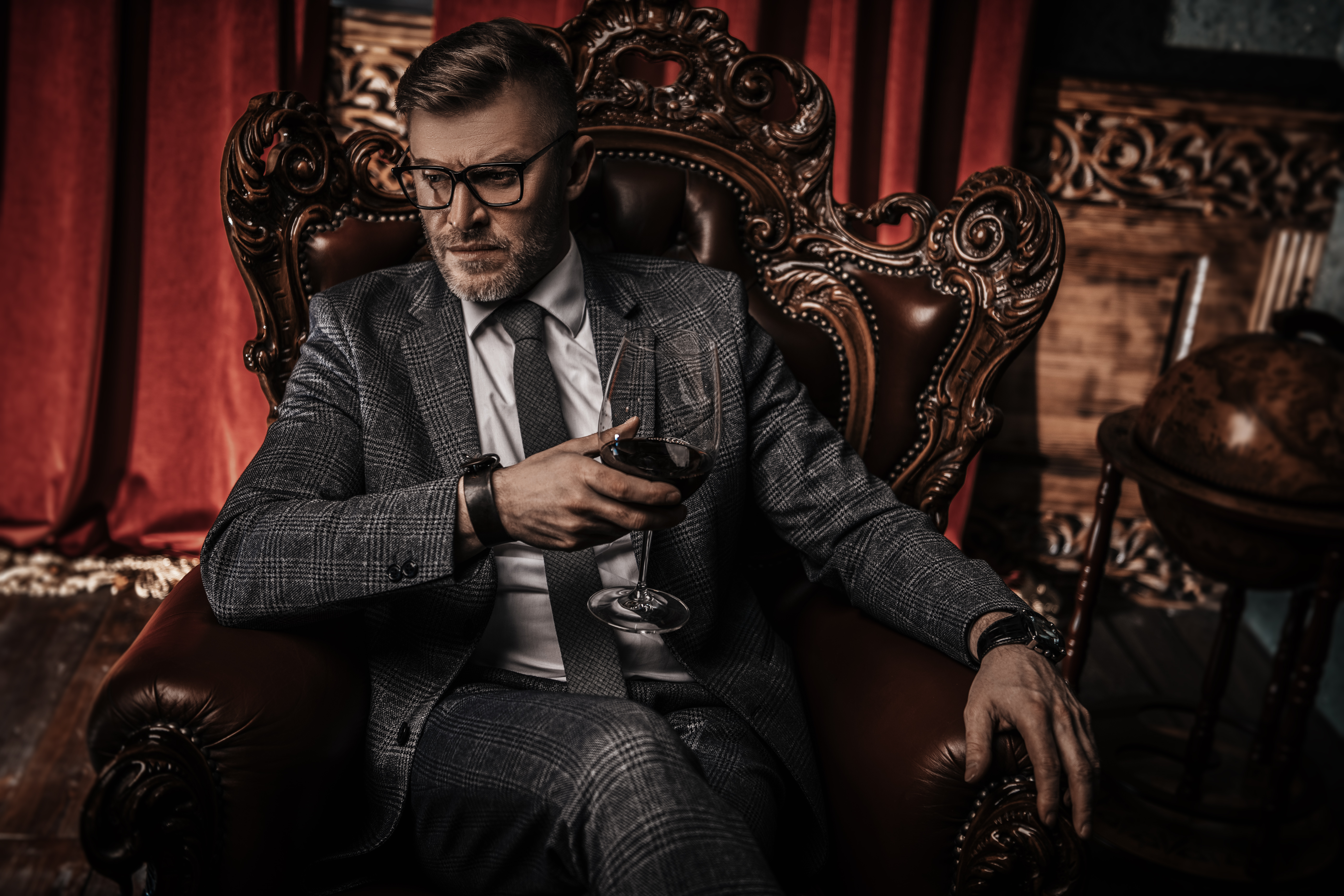 Retrato de un apuesto hombre maduro con traje formal bebiendo vino en el sillón de un interior clásico. | Fuente: Shutterstock