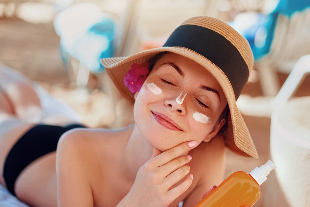 Mujer joven con crema del sol en la cara sostiendo la botella de bloqueador solar en la playa. | Fuente: Shutterstock