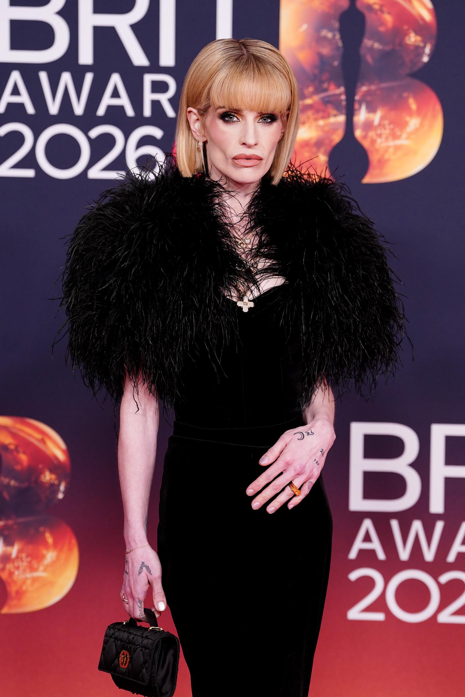 Kelly Osbourne lució un vestido negro ceñido con unos espectaculares hombros adornados con plumas, un collar con una cruz y un bolso de mano en miniatura en los BRIT Awards de 2026, celebrados en Mánchester. | Fuente: Getty Images.