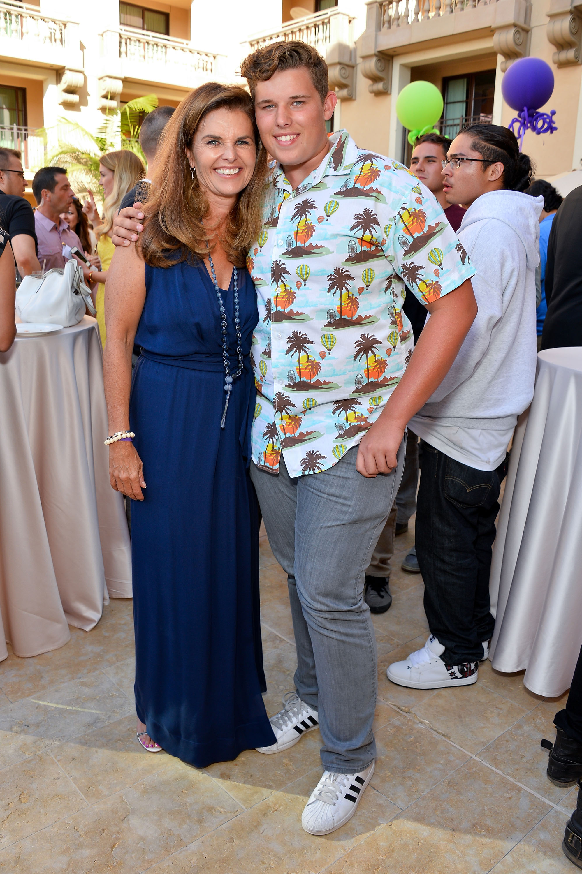 Maria Shriver y su hijo Christopher comparten un cálido momento en el mismo evento. Shriver lleva un vestido azul oscuro con un collar, mientras que Christopher luce su característica camisa estampada y vaqueros grises. Están muy juntos, rodeados de invitados en un ambiente festivo.