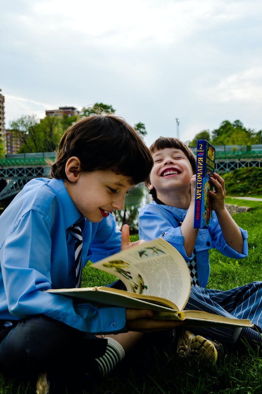 Dos niños sentados en la hierba mientras ríen y leen libros | Foto: Pexels