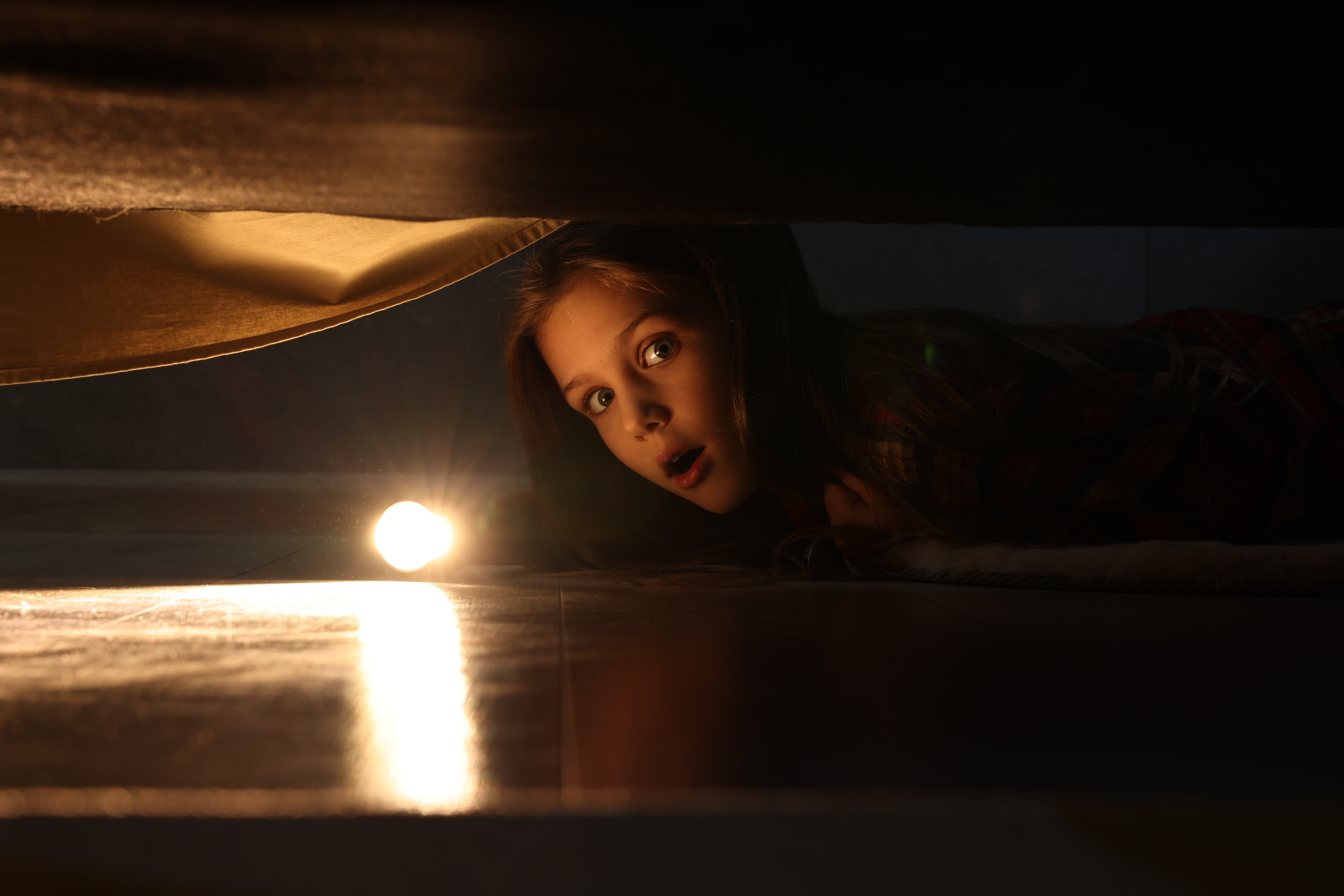 Una niña sorprendida sujetando una linterna y mirando debajo de la cama | Foto: Shutterstock