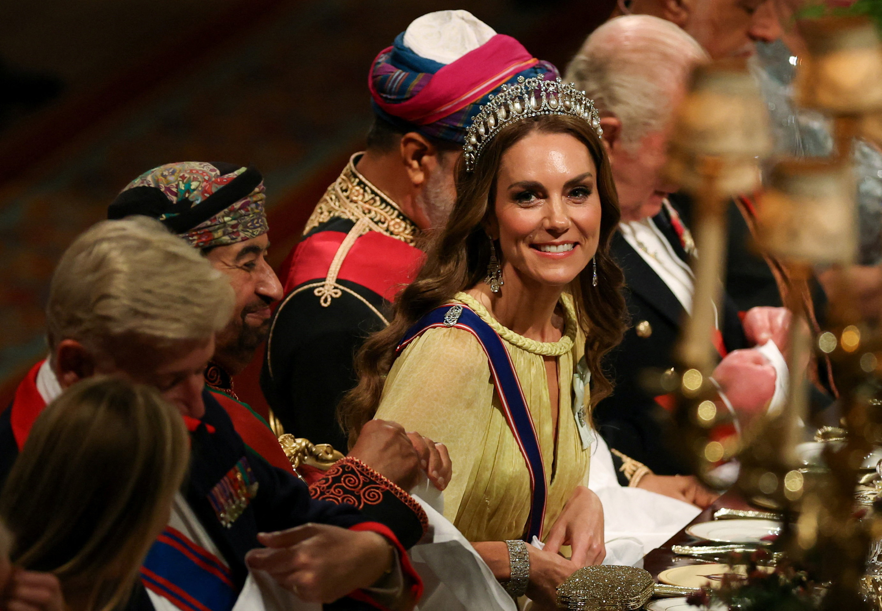 Sentada a la mesa del banquete en el salón de San Jorge el 20 de octubre de 2026, Catherine, princesa de Gales, sonríe al comenzar la cena. Luce ondas castañas sobre los hombros bajo la tiara Lover's Knot de perlas y diamantes, pendientes largos de diamantes, maquillaje de ojos ahumados y un vestido amarillo con capa y escote de cordón trenzado. | Fuente: Getty Images