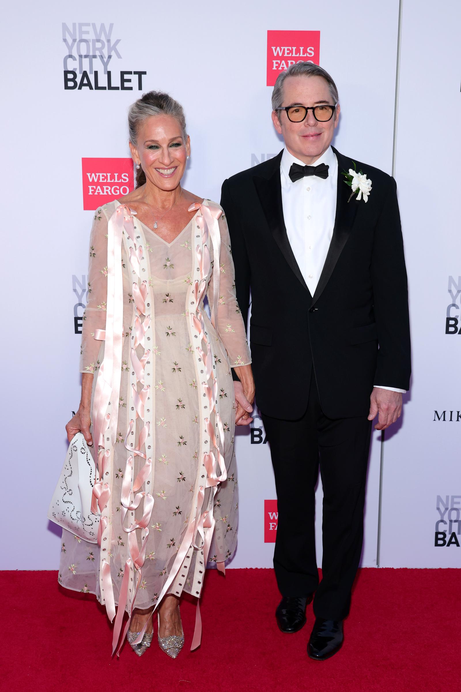 Sarah Jessica Parker va de la mano de Matthew Broderick mientras se puede ver en todo su esplendor su vestido floral semitransparente, con cintas rosas que le caen por la cola, unos zapatos de tacón brillantes y un bolso blanco. | Fuente: Getty Images