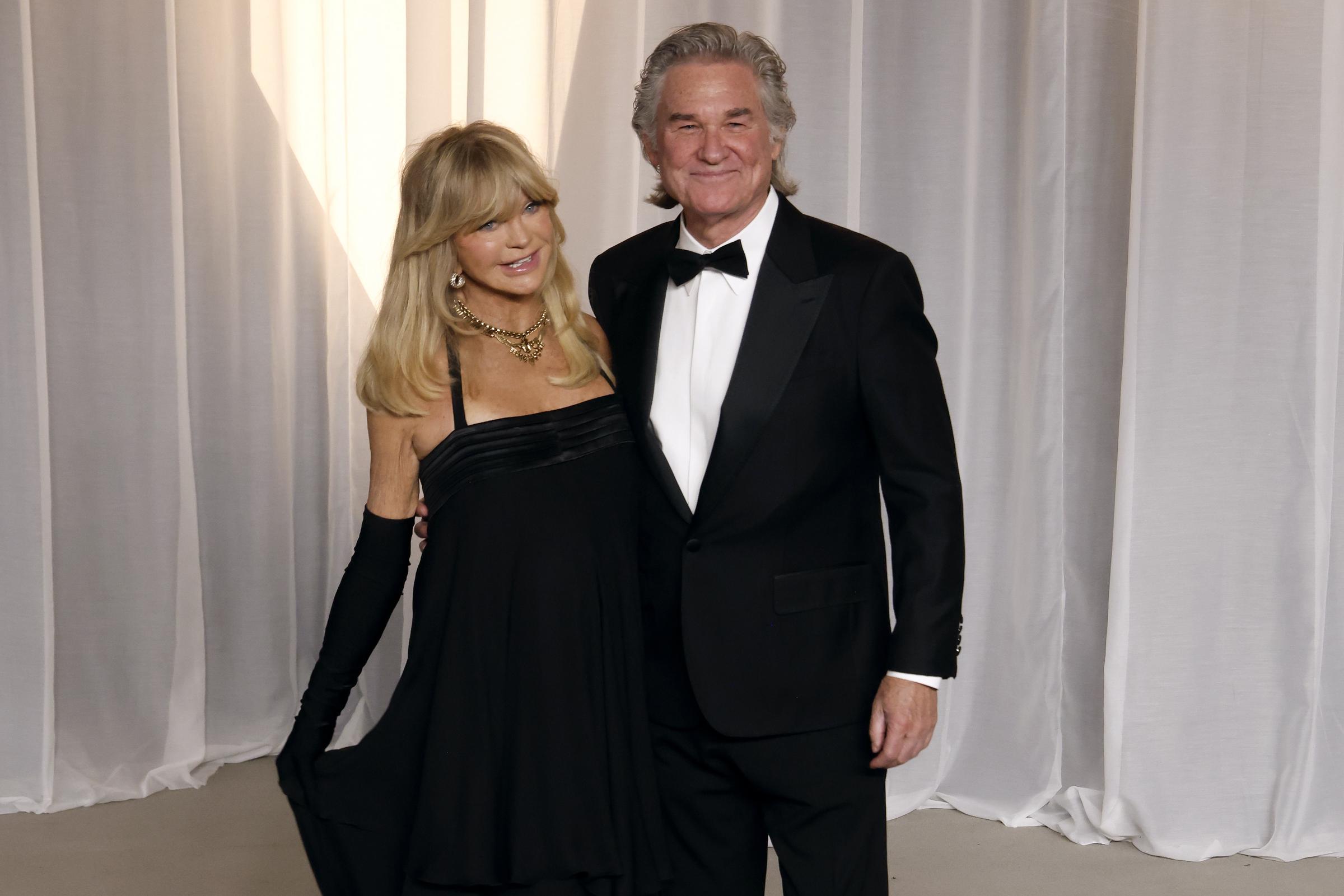 Goldie Hawn y Kurt Russell forman una pareja elegante y natural con sus trajes negros de gala a juego. Hawn está encantadora con un vestido de cóctel negro de corte imperio, combinado con guantes largos a juego, llamativas joyas de oro y su característico flequillo rubio y voluminoso. Russell complementa su estilo clásico con un elegante esmoquin negro con solapas en punta de satén, una camisa blanca impecable y un corbatín negro tradicional.