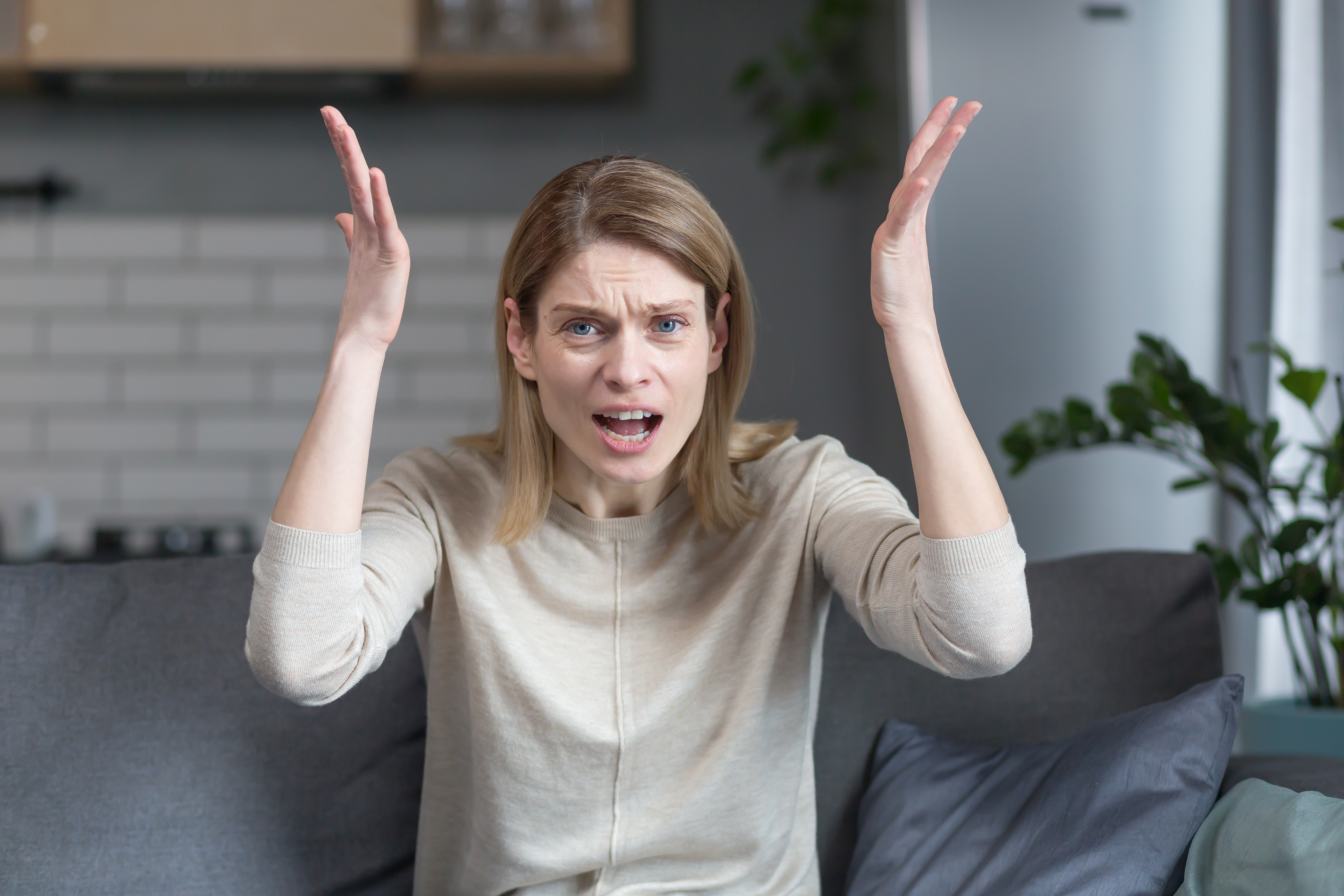 Mujer enfadada y frustrada hablando por videollamada mirando a la webcam, gesticulando airadamente con las manos mientras está sentada en el sofá de su casa | Foto: Getty Images