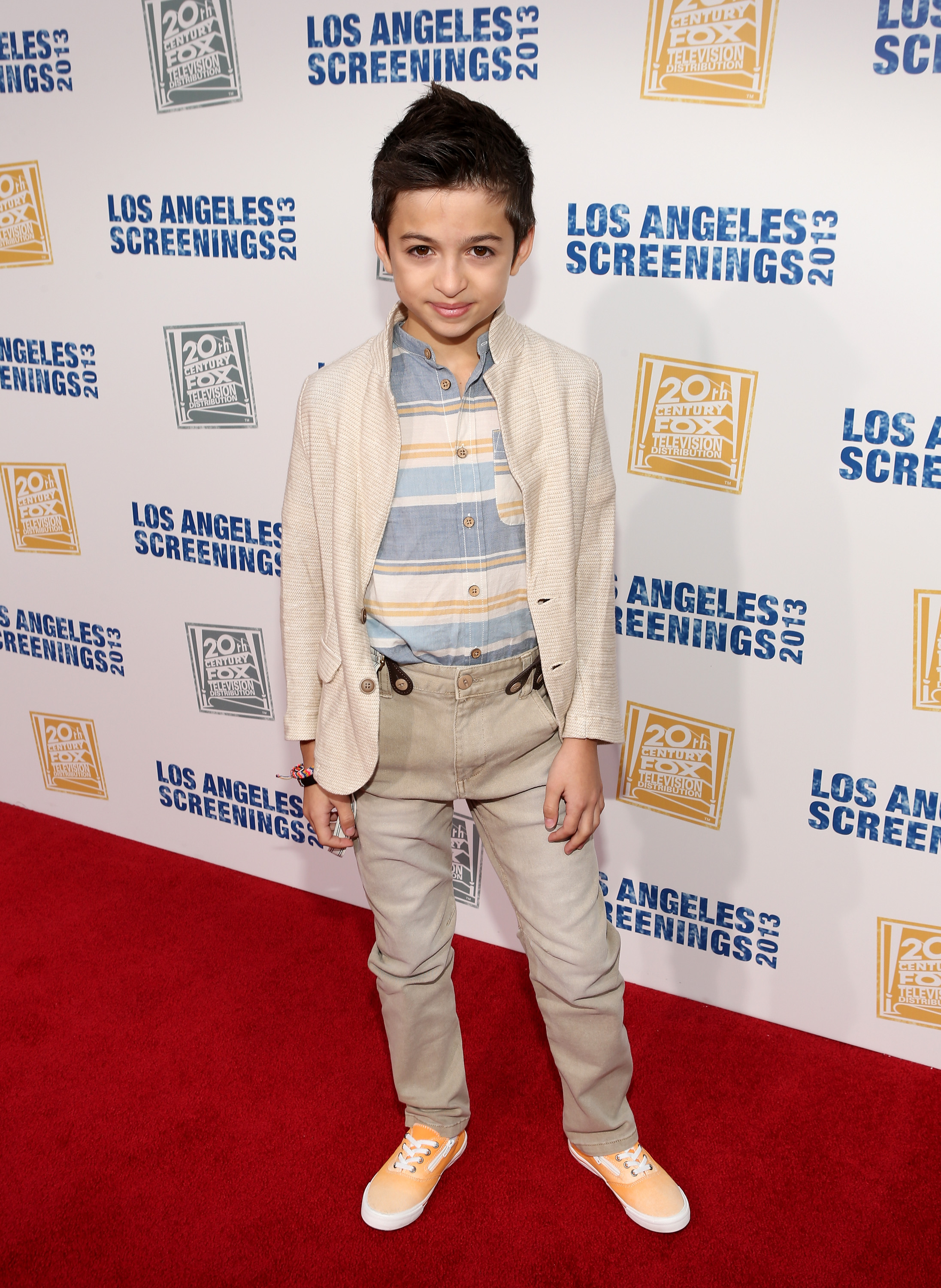 Posando en la alfombra roja de la fiesta "LA Screenings Lot Party" de Twentieth Century Fox Television Distribution de 2013, celebrada en Los Ángeles, California, con una chaqueta color crema, una camisa a rayas, pantalones beige y zapatillas naranjas. | Fuente: Getty Images