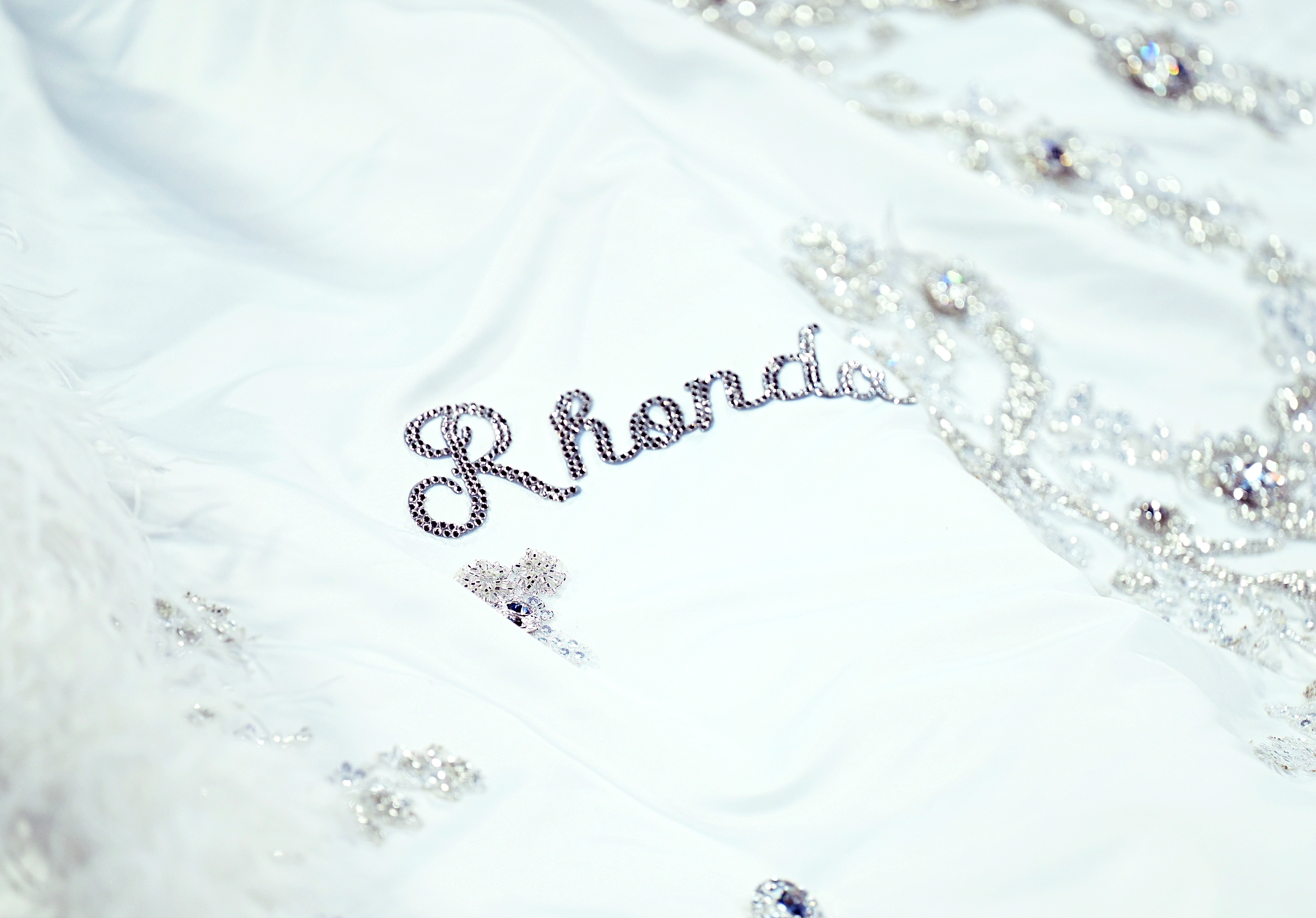 Un primer plano de la cola resalta el bordado exquisito, con el nombre “Rhonda” claramente visible entre los delicados detalles. Los demás nombres, de sus hijos y nietos, se asoman sutilmente, ofreciendo destellos que realzan el emotivo diseño del vestido.