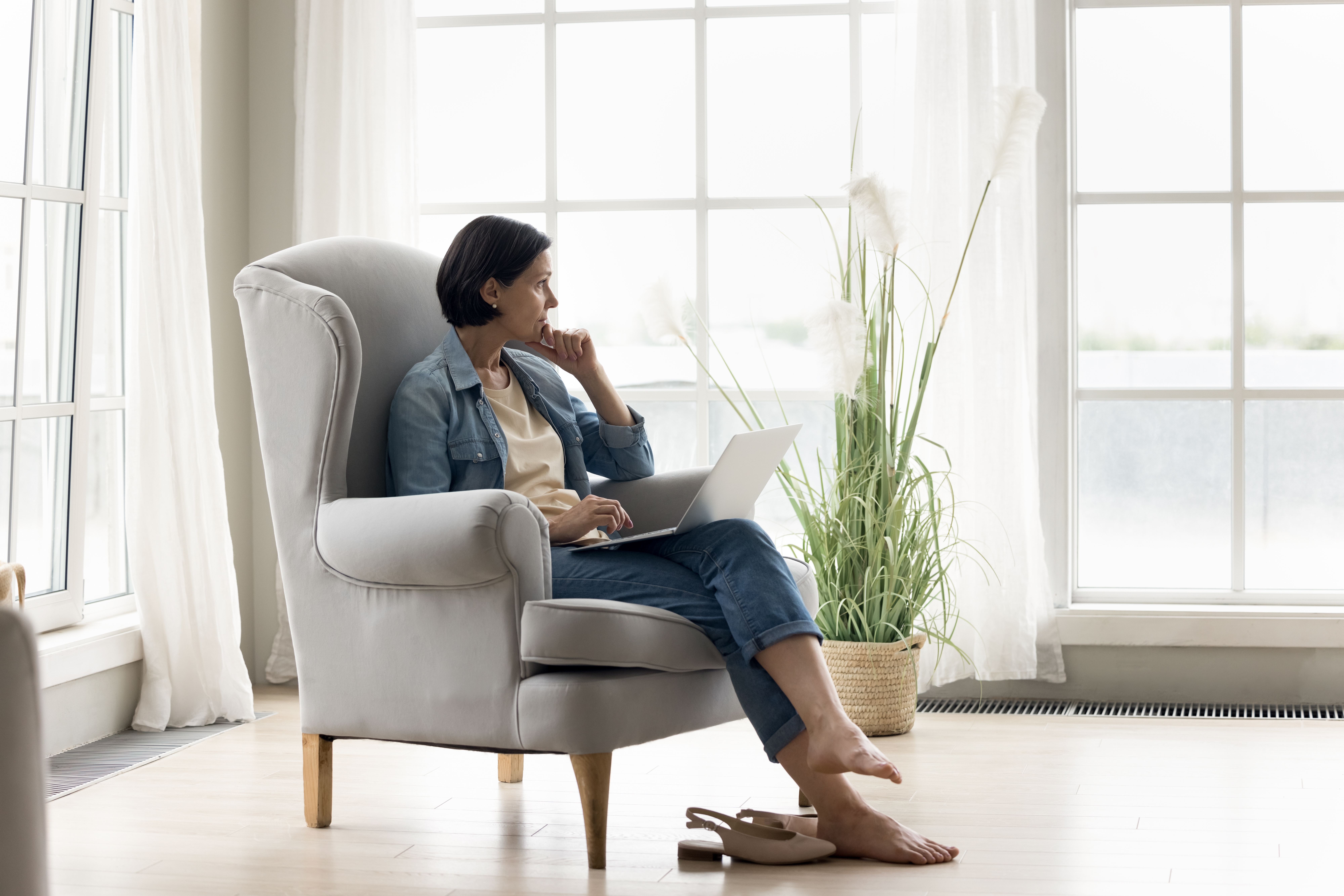 Mujer mirando por la ventana sentada en una silla con su portátil | Foto: Shutterstock