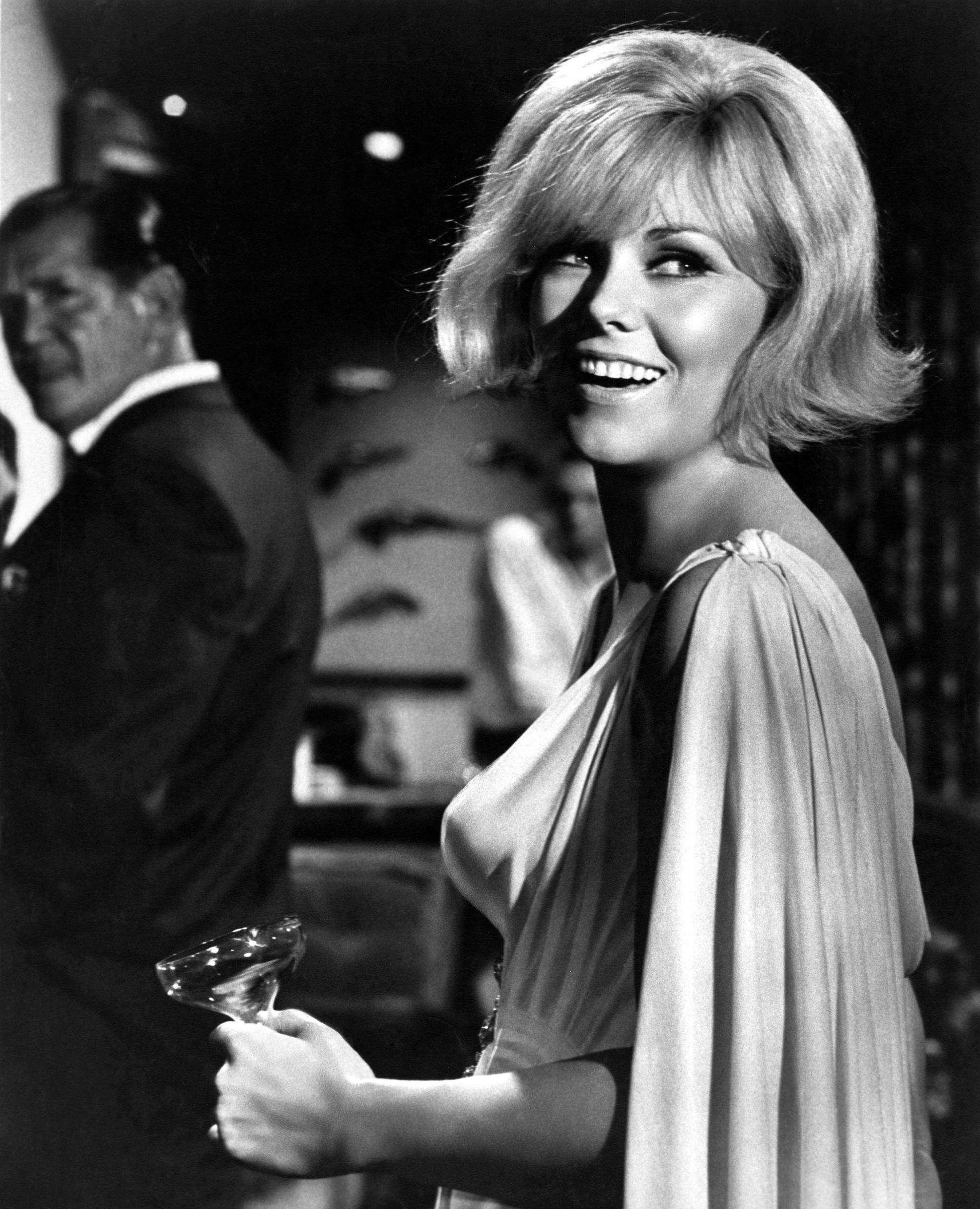 Kim Novak sonríe mientras sostiene una copa de cóctel en el set de filmación de The Legend of Lylah Clare, luciendo un fluido vestido de noche. | Fuente: Getty Images