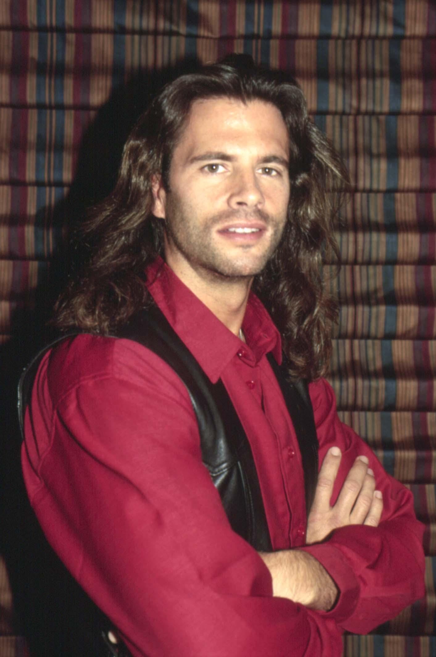 Lorenzo Lamas evoca su ruda imagen de "Renegade" en un retrato de 1993. Con los brazos cruzados ante un fondo a rayas, el actor luce cabello oscuro ondulado hasta los hombros, barba incipiente y su característica mirada intensa. Viste camisa roja oscura abotonada y chaleco de cuero negro. | Fuente: Getty Images