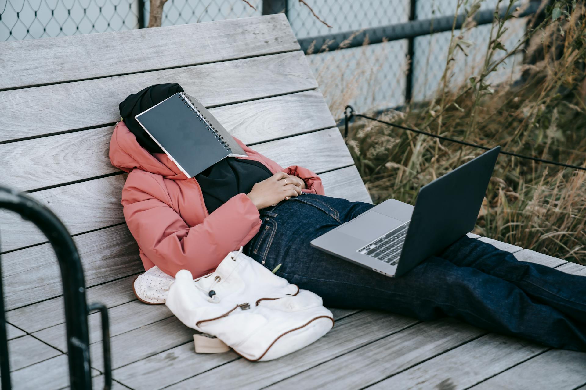 Una mujer irreconocible con un portátil abierto descansando en un banco con un libro en la cara | Fuente: Pexels