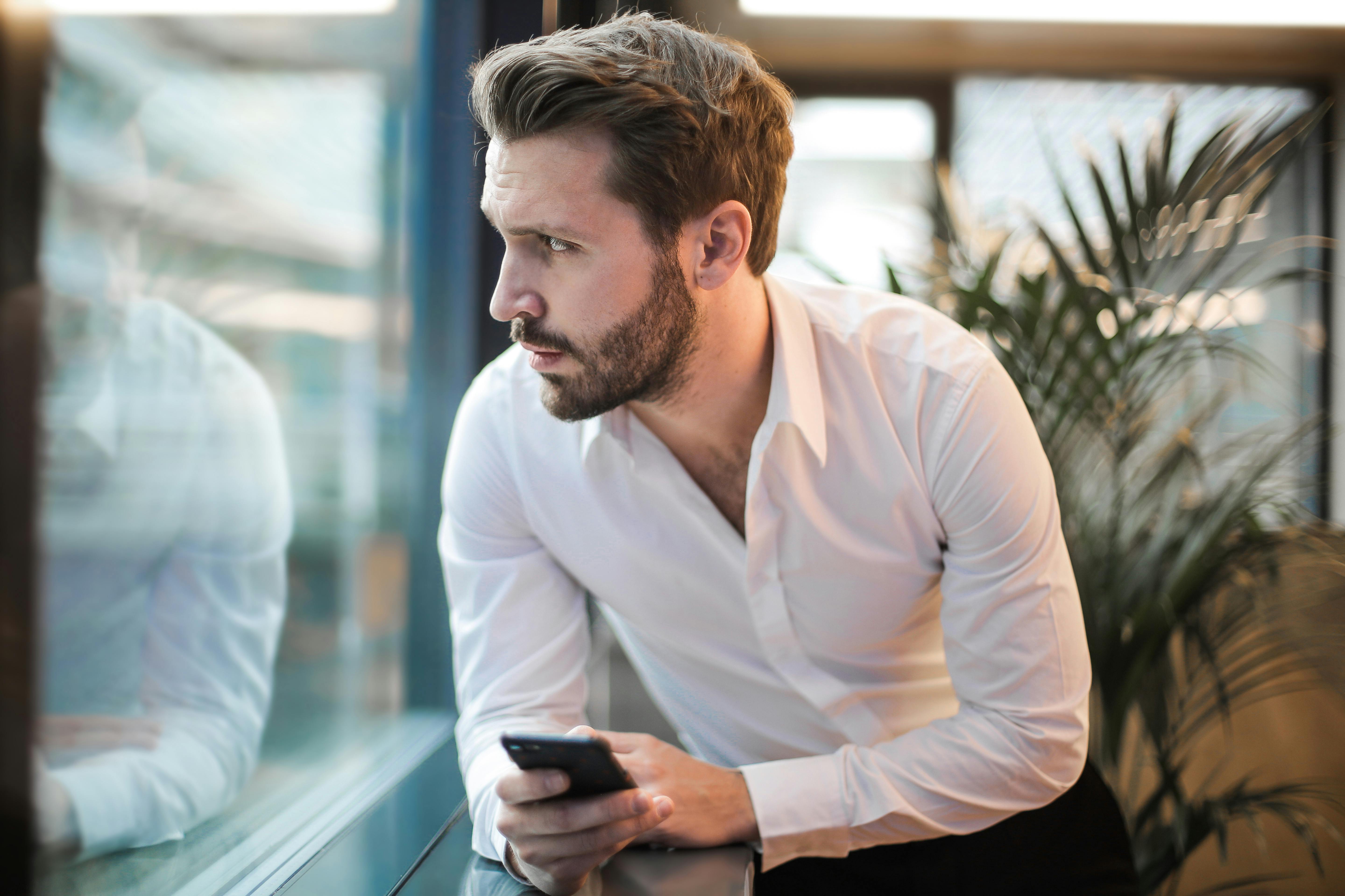 Un hombre mirando por la ventana, teléfono en mano | Fuente: Pexels