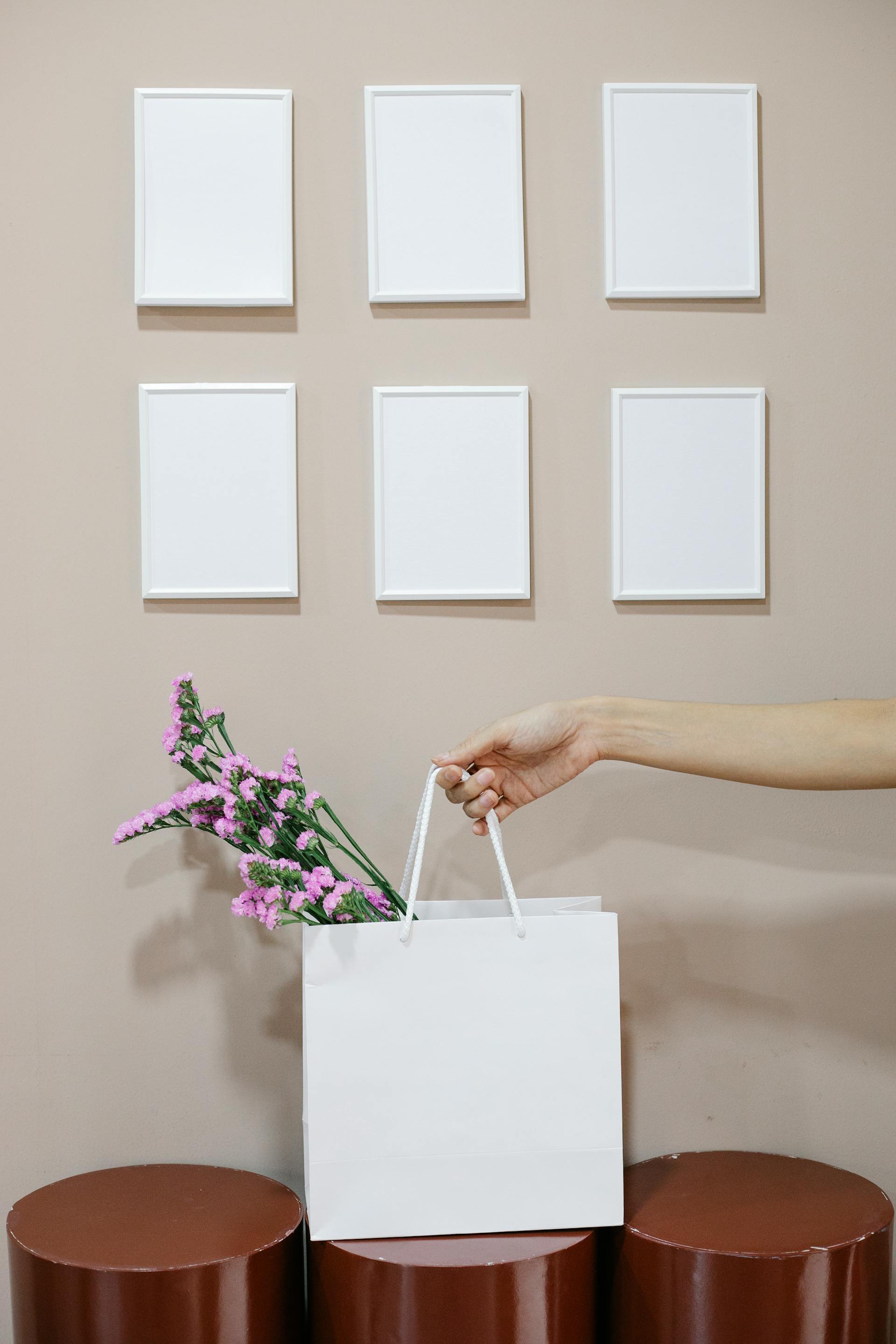 Foto en primer plano de una mujer que sostiene una bolsa de papel con flores cerca de una pared con marcos en blanco | Foto: Pexels
