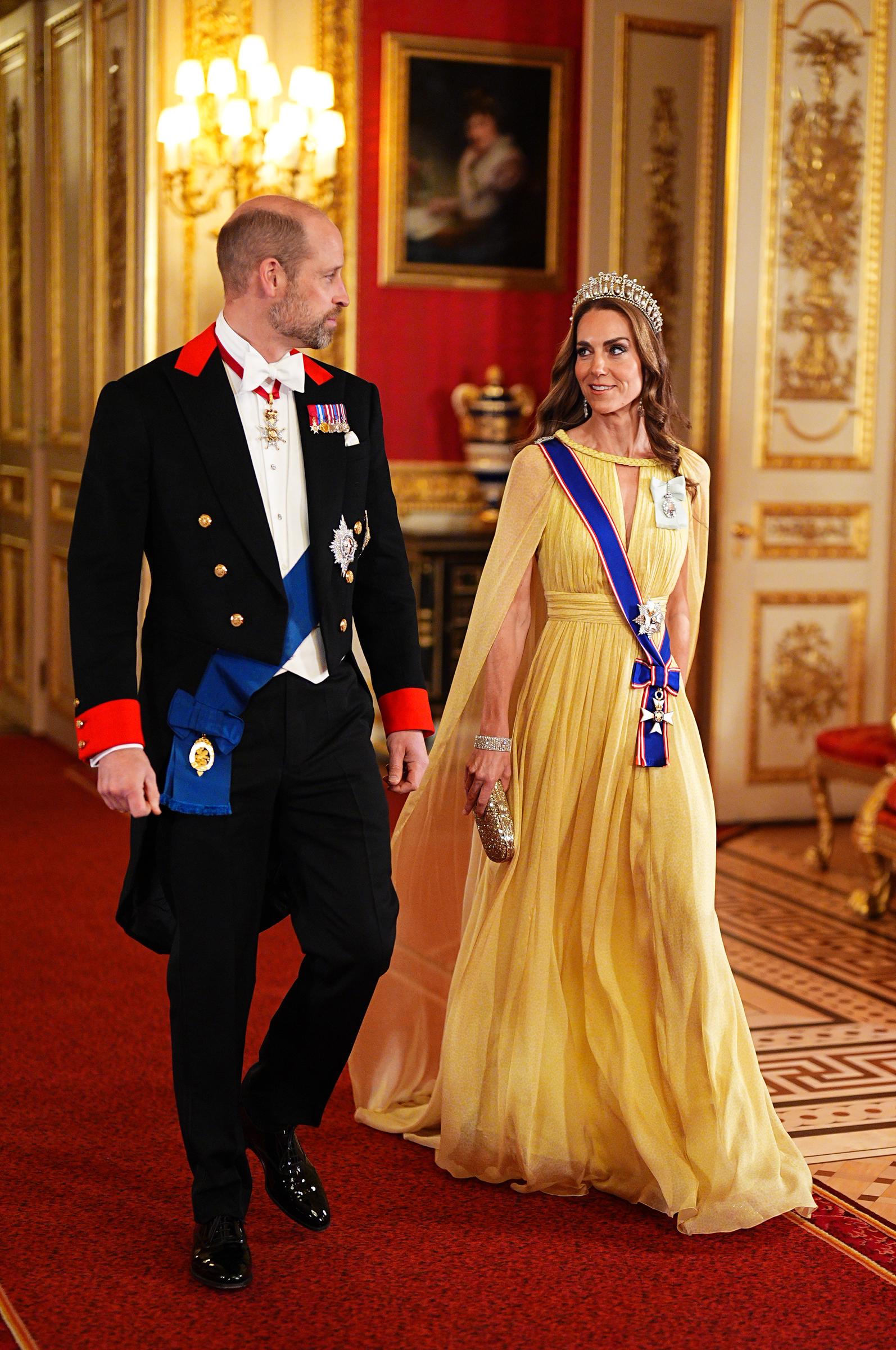 Los príncipes de Gales caminan juntos por los pasillos alfombrados de rojo del castillo de Windsor el 20 de octubre de 2026, luciendo elegantes antes de la cena de gala. Catherine viste un traje amarillo pastel plisado con capa de Elie Saab y lleva un brillante bolso de mano dorado. El príncipe William luce frac entallado, chaleco blanco, moño y las insignias de la Orden de la Jarretera. | Fuente: Getty Images