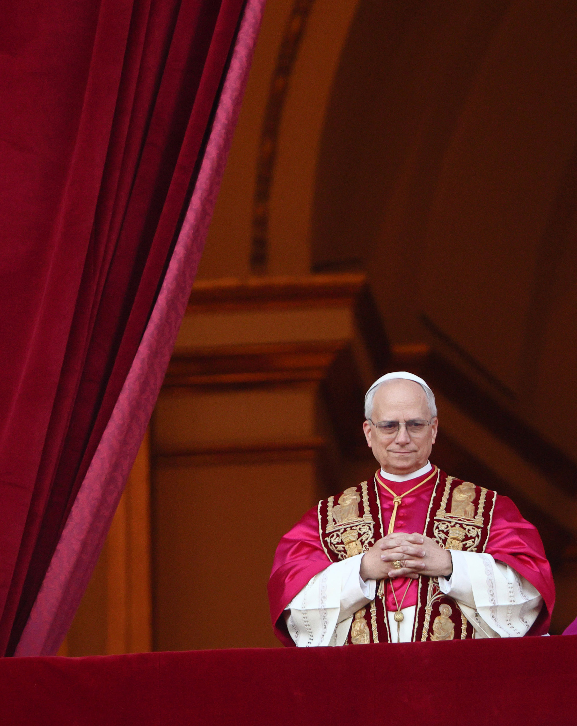 Pope Leo XIV stands solemnly between rich velvet drapes, hands clasped over ornate crimson vestments embroidered with golden saints