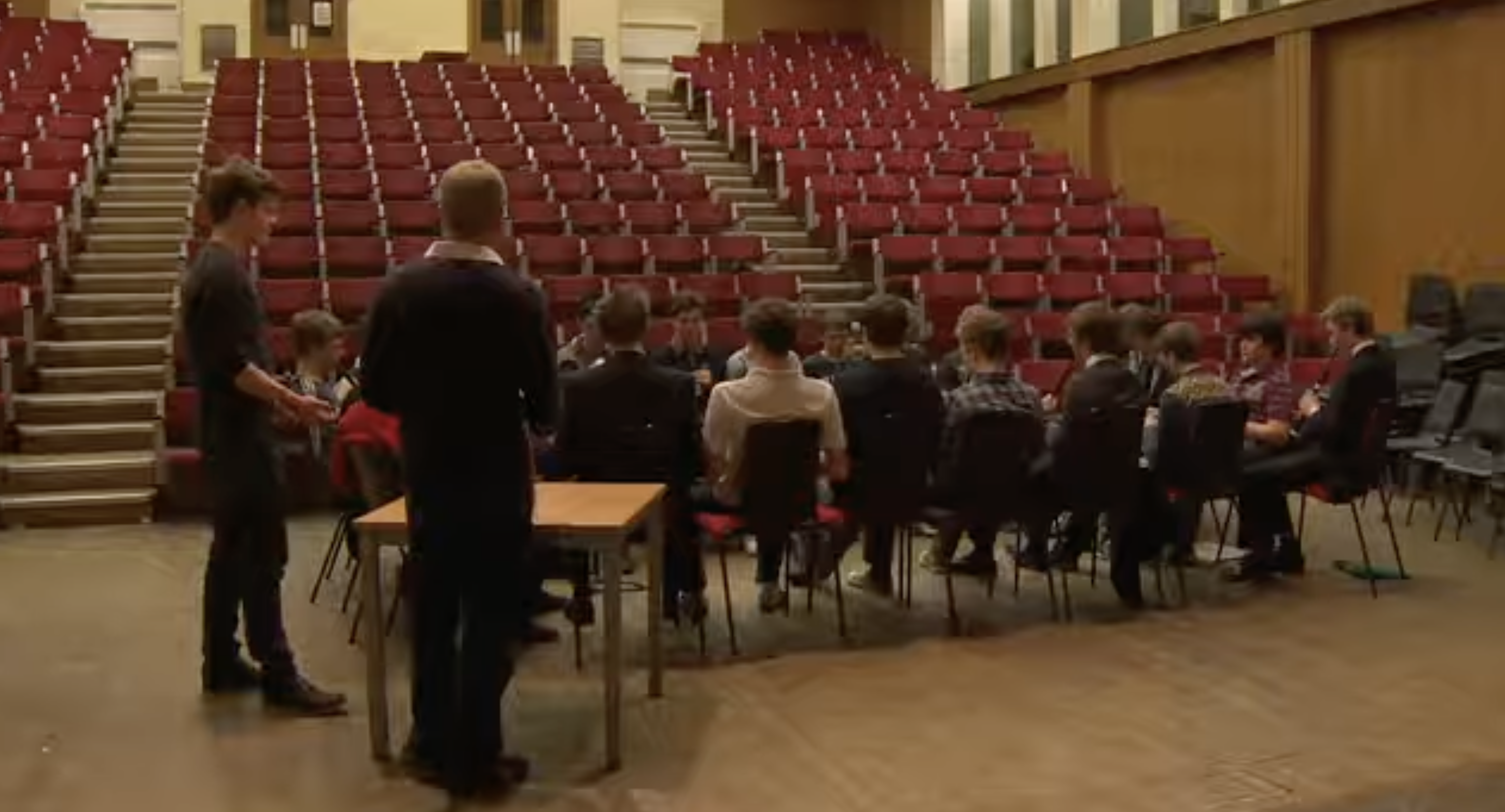 Los alumnos están sentados en grupo en el suelo de un gran salón rodeado de filas de gradas rojas. La imagen permite echar un vistazo a los espacios comunes que se utilizan para las actividades en el Eton College. | Fuente: YouTube/Paradoxyy
