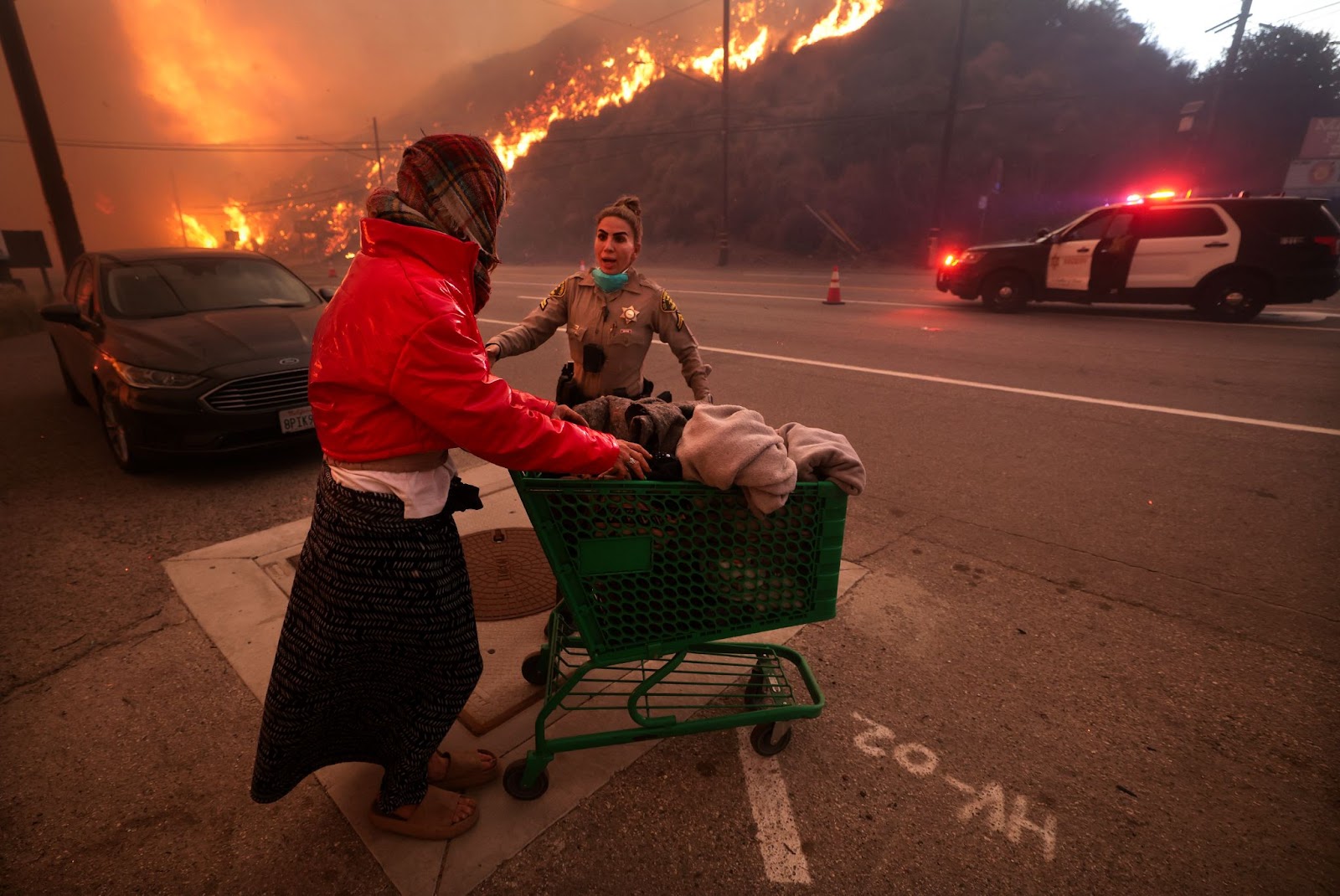 Un agente de policía escolta a una mujer sin hogar lejos de Pacific Coast Highway y Topanga Canyon Boulevard mientras el incendio de Palisades arrasa las colinas en Los Ángeles, California, el martes 7 de enero de 2025. | Fuente: Getty Images