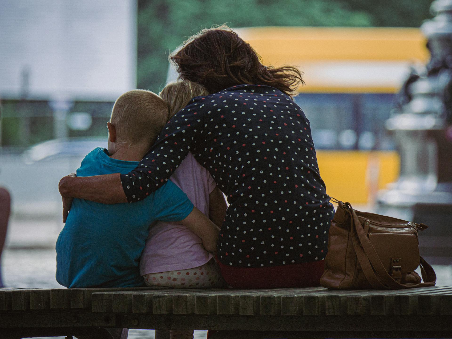 Toma de espaldas de una mujer abrazando a sus hijos sentada en un banco | Fuente: Pexels
