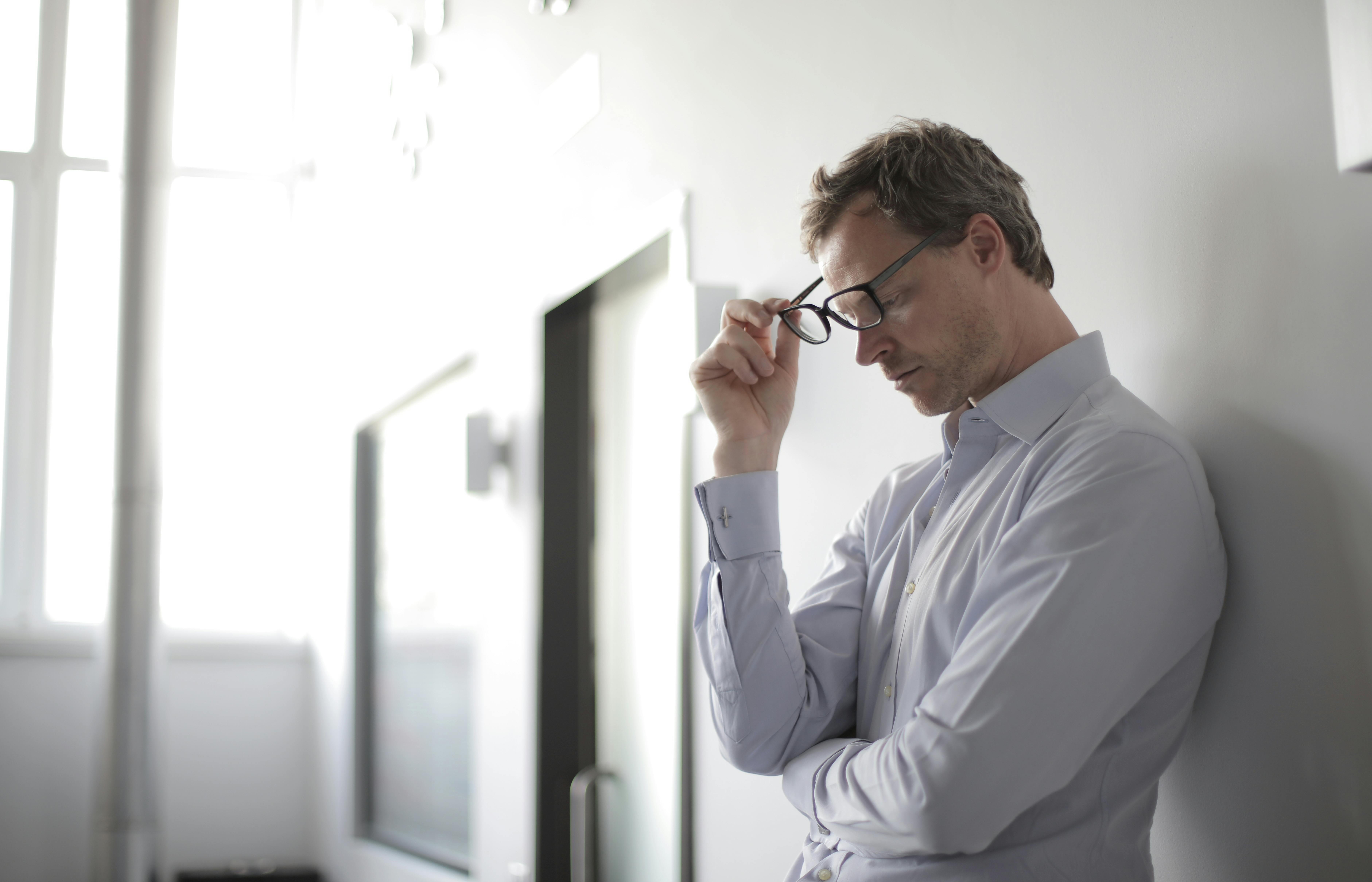 Un hombre alterado apoyado en una pared y quitándose las gafas | Fuente: Pexels
