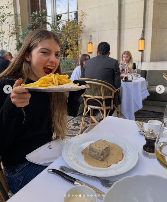 Con gesto divertido y la boca abierta a punto de comer, Antonia Macri sostiene una bandeja colmada de papas fritas doradas frente a un plato principal con salsa servido en la terraza de un restaurante. | Fuente: Instagram/juliana.awada