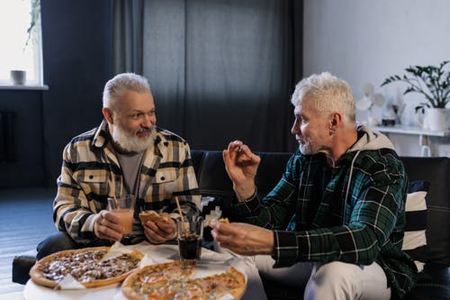 Dos hombres mayores conversan comiendo pizza. | Foto: Pexels