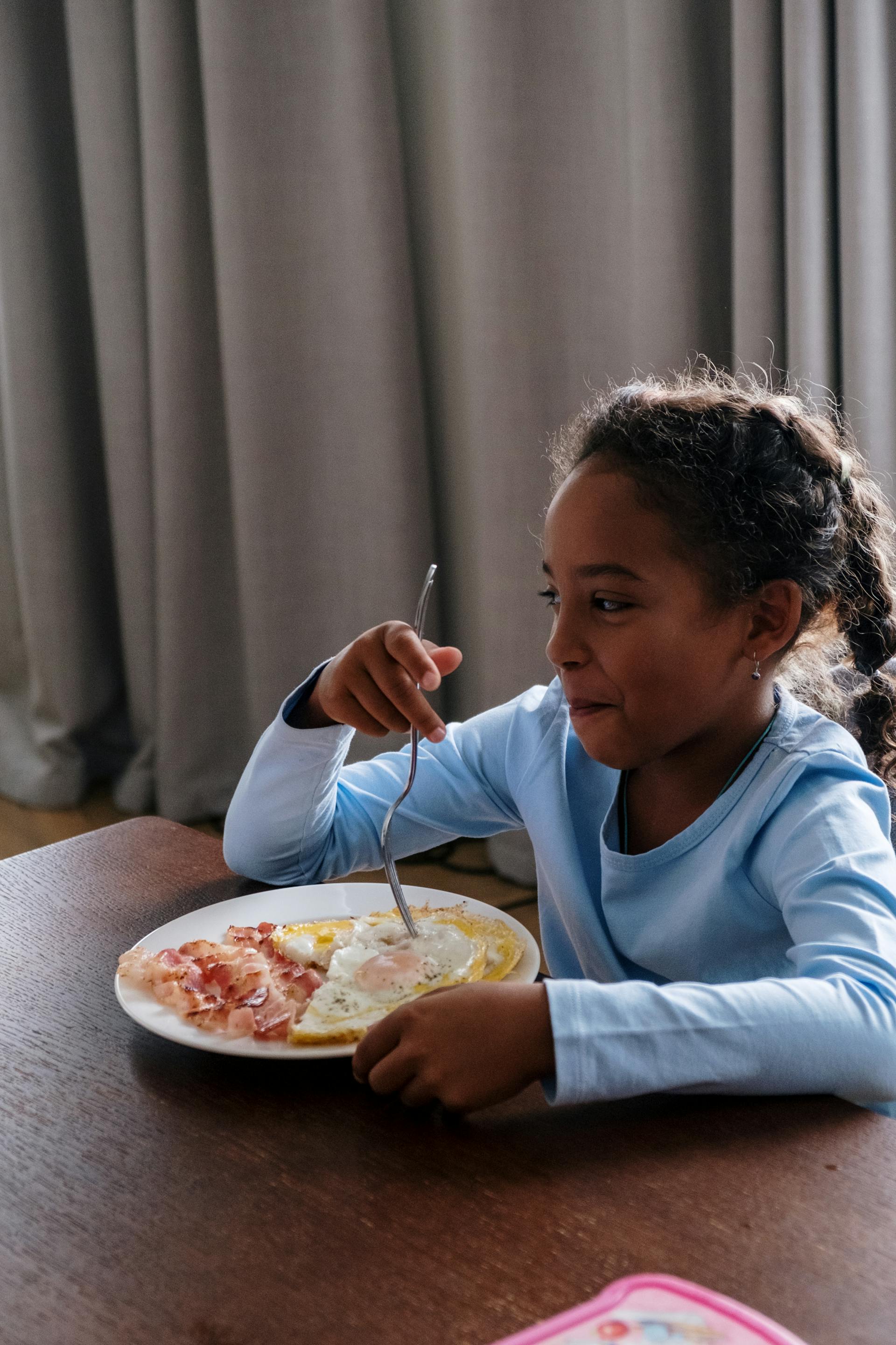 Una niña desayunando intenta reprimir la risa mientras mira a alguien | Fuente: Pexels