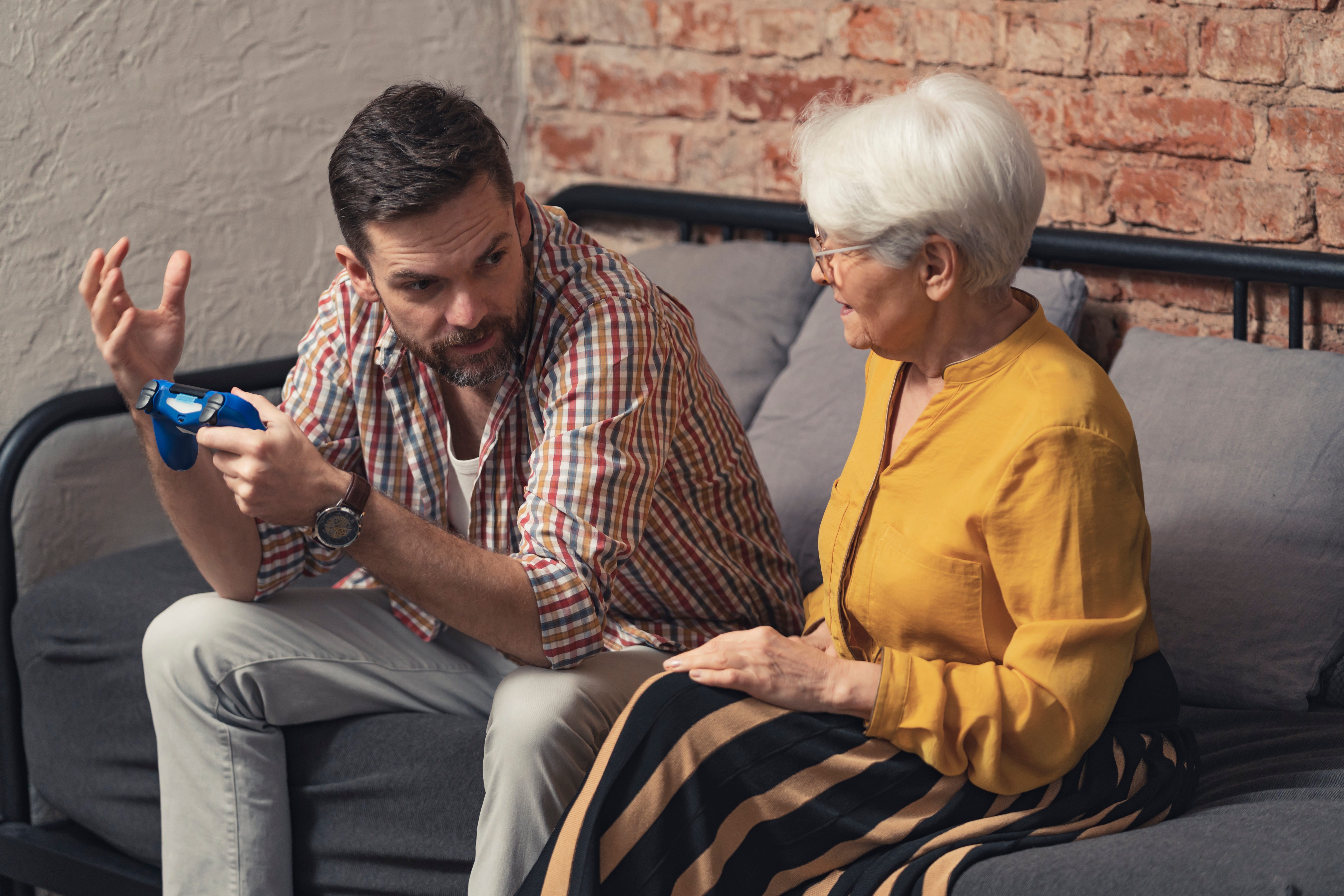 Un hombre sentado en un sofá sujetando un mando de videojuegos mientras gesticula con las manos a una señora mayor. | Foto: Shutterstock
