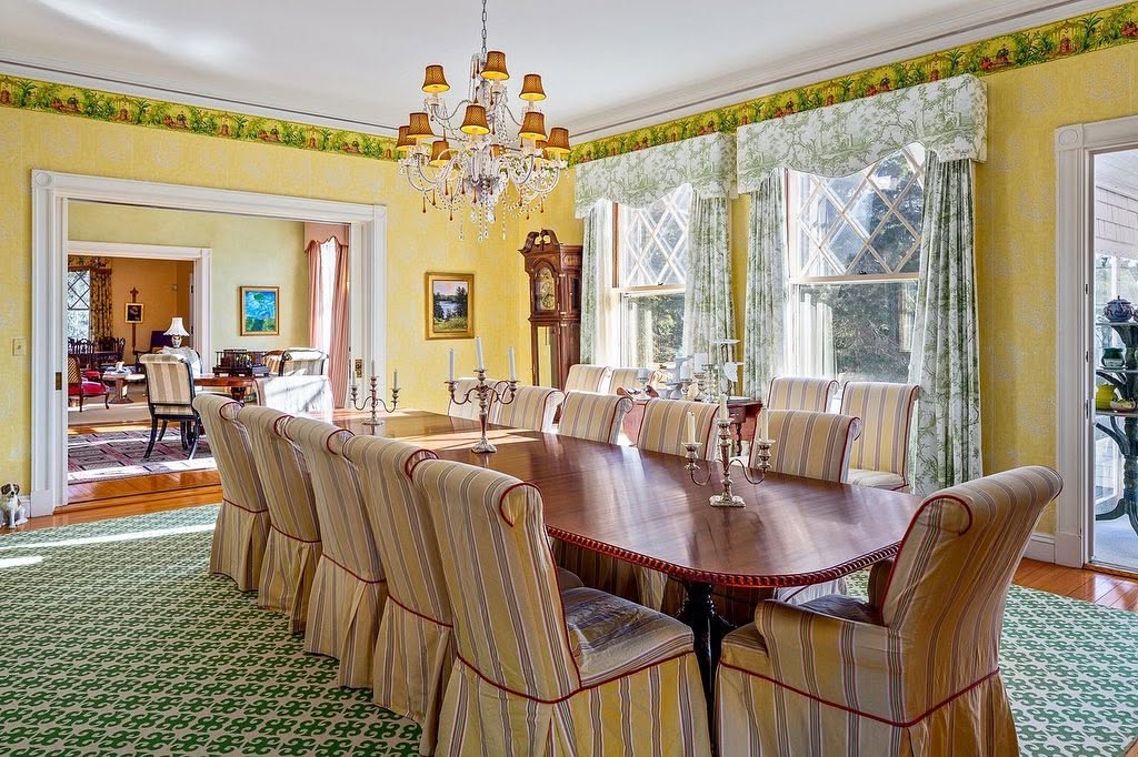 El comedor formal cuenta con una mesa larga de madera, sillas a rayas, una lámpara de araña de cristal y ventanas con cristales en forma de rombo que dan a la finca. | Fuente: Facebook/LegacySIR
