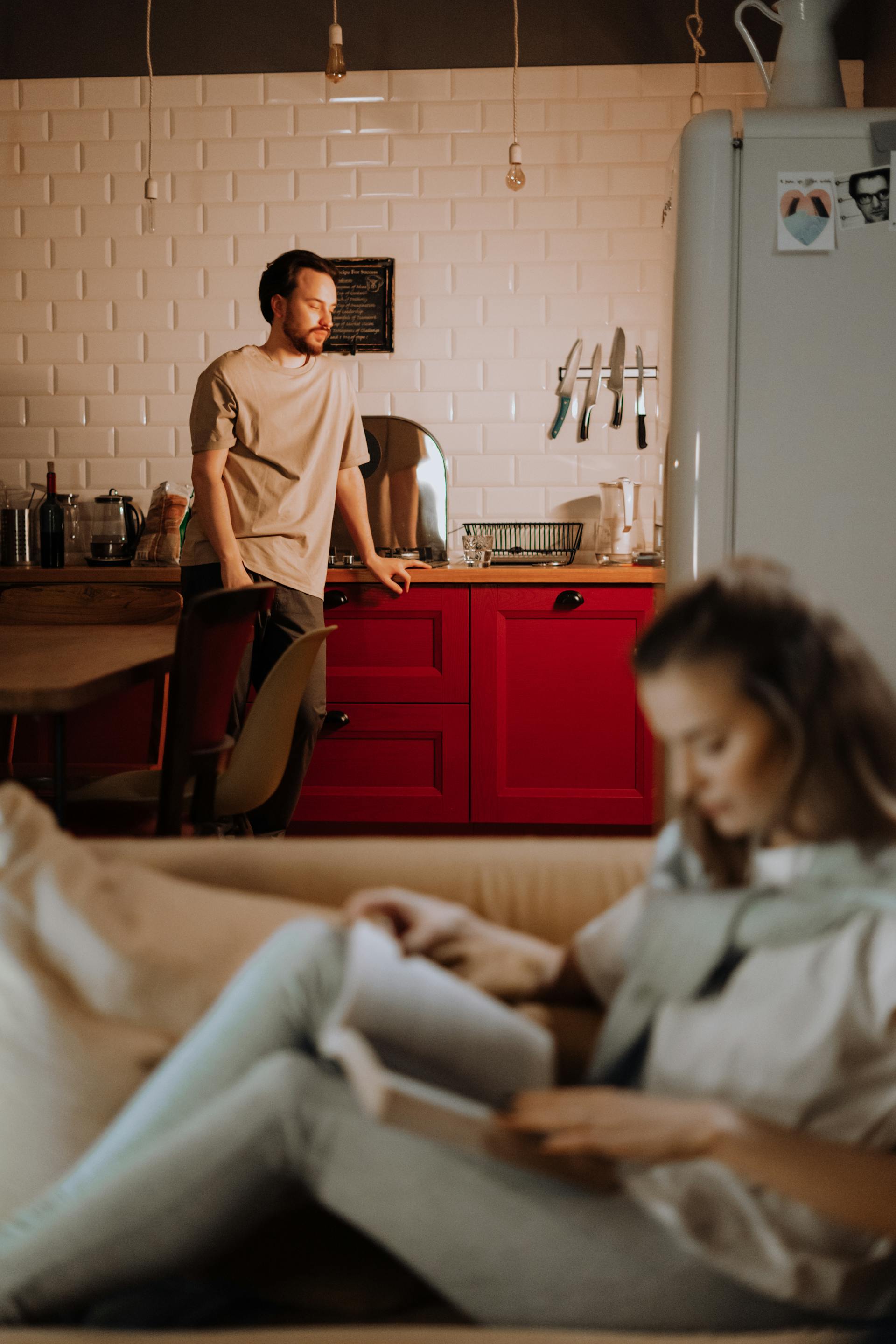 Una mujer lee un libro en el sofá mientras un hombre angustiado está de pie en la cocina | Fuente: Pexels