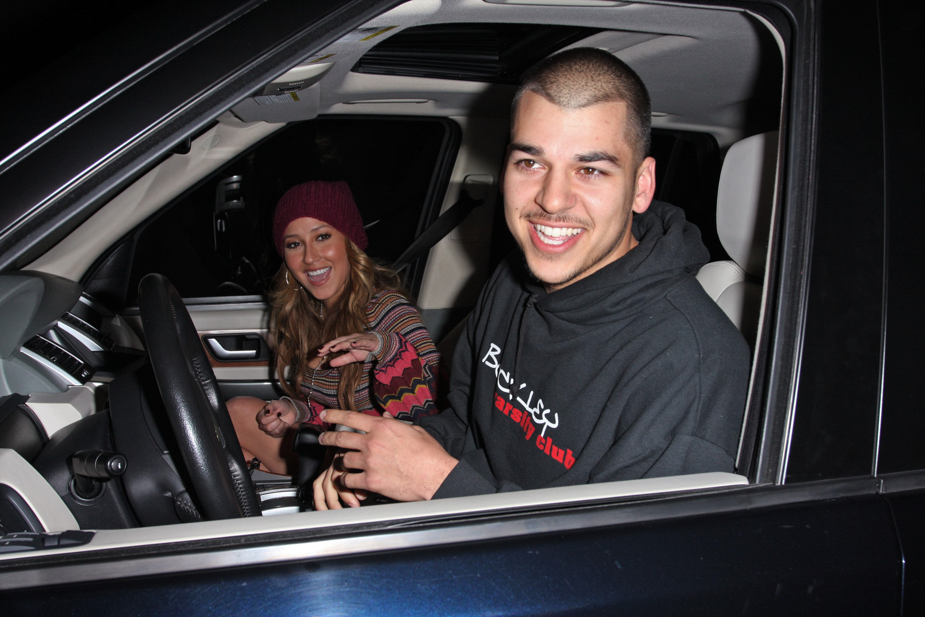 Rob Kardashian sonríe al volante mientras una mujer sentada a su lado saluda a los fotógrafos durante una salida nocturna. La foto capta un momento relajado y alegre de sus primeros años bajo los focos. | Fuente: Getty Images.