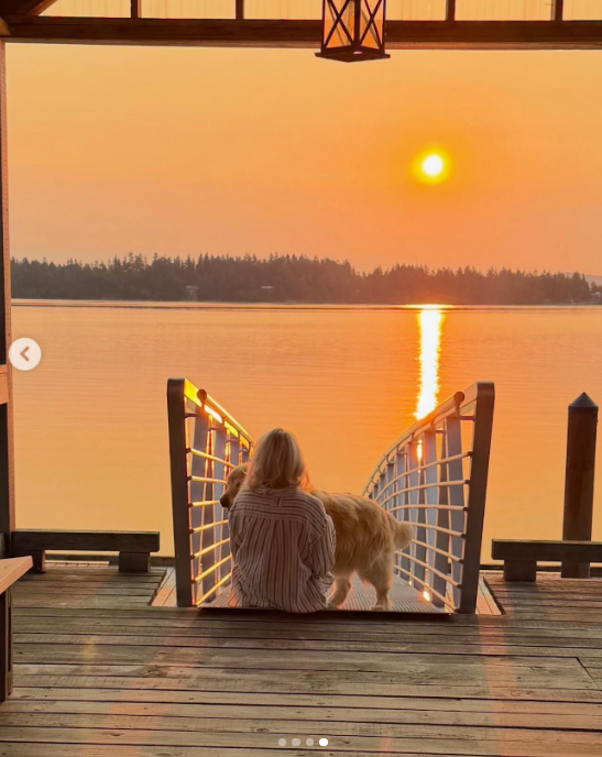 Pamela Anderson disfruta de los ritmos tranquilos de la vida costera en Arcady, sentada junto a uno de sus perros en un muelle de madera durante una radiante puesta de sol en Ladysmith, Columbia Británica. Vista de espaldas, con una camisa a rayas oversize abotonada, contempla el agua resplandeciente mientras el sol del atardecer se esconde tras la línea de árboles, y comparte este momento de serenidad con sus seguidores junto a la leyenda: "Una chica isleña de corazón 🤍". | Fuente: Instagram/pamelaanderson