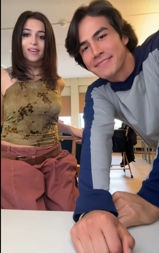 Josie Totah sale en un vídeo de TikTok con un chico, sentada a su lado en una mesa en lo que parece ser un local de ensayo o un estudio. Lleva un top floral verde oliva, pantalones cargo rosa empolvado y varios collares superpuestos, mientras que su acompañante lleva una camisa de manga larga gris y azul marino. | Fuente: TikTok/josie totah
