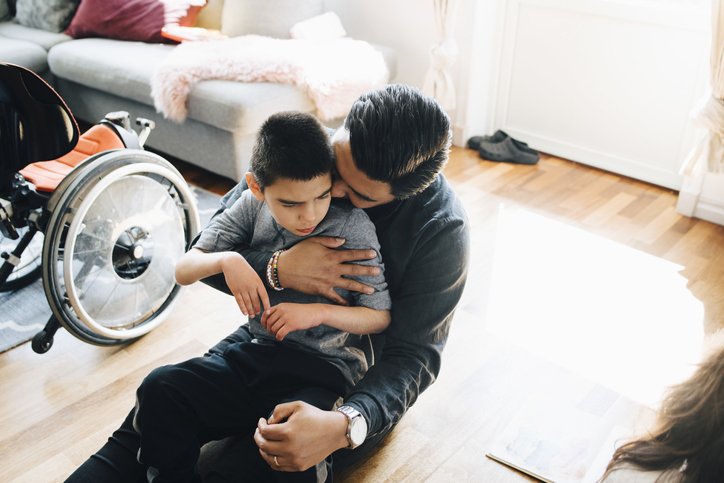 Padre sentado en el piso abraza a su hijo con parálisis cerebral. | Foto: Shutterstock
