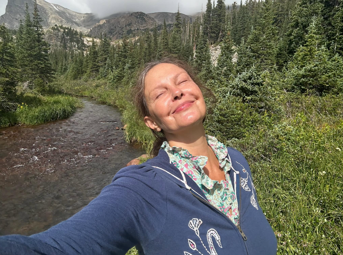 Judd cierra los ojos y sonríe bajo el sol durante una excursión a la montaña en 2025. Con una sudadera azul con capucha junto a un arroyo, disfruta de un momento de relajación lejos de la alfombra roja. | Fuente: Facebook/AshleyJuddOfficial
