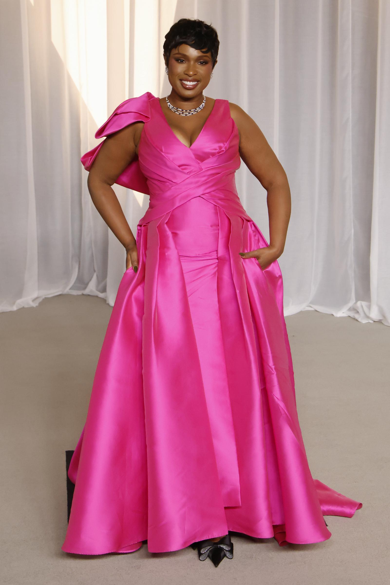 Jennifer Hudson acapara todas las miradas con un vestido de satén rosa intenso y llamativo, con un escote en V muy pronunciado y drapeados esculpidos a lo largo del corpiño. Un lazo asimétrico y voluminoso resalta uno de los hombros, mientras que la falda amplia, que roza el suelo, incluye bolsillos integrados que le dan un toque de encanto natural. La ganadora del EGOT completa este look espectacular con un brillante collar de diamantes, unos zapatos de tacón de punta adornados con lazos de cristal y un elegante corte de pelo pixie.