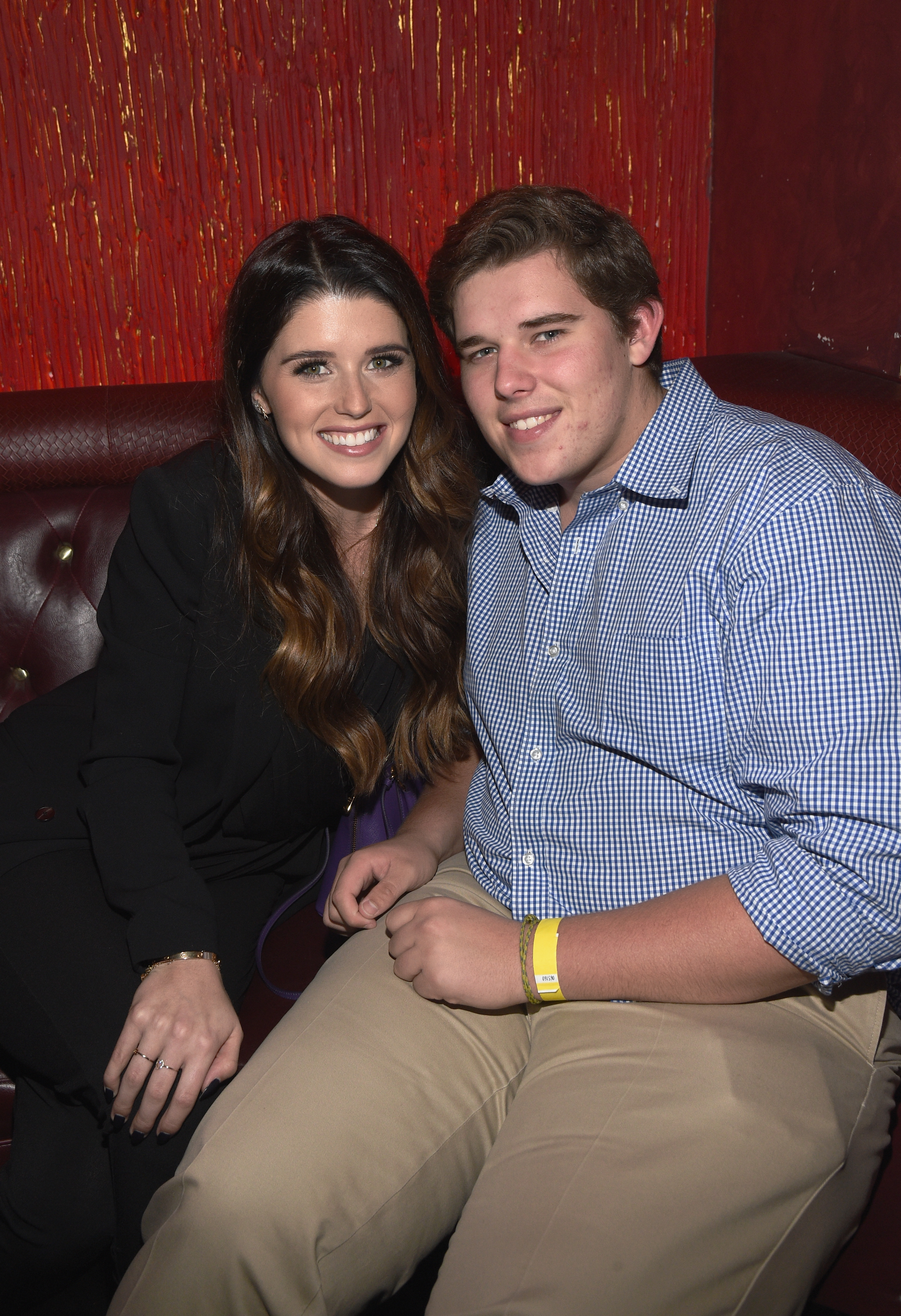 Christopher sentado junto a su hermana Katherine en la recepción previa a un evento en Los Ángeles, en diciembre de 2014. Lleva una camisa azul a cuadros y pantalones beige, con una muñequera amarilla visible en la muñeca derecha. Su rostro más redondeado y su actitud relajada reflejan su estilo durante este periodo.