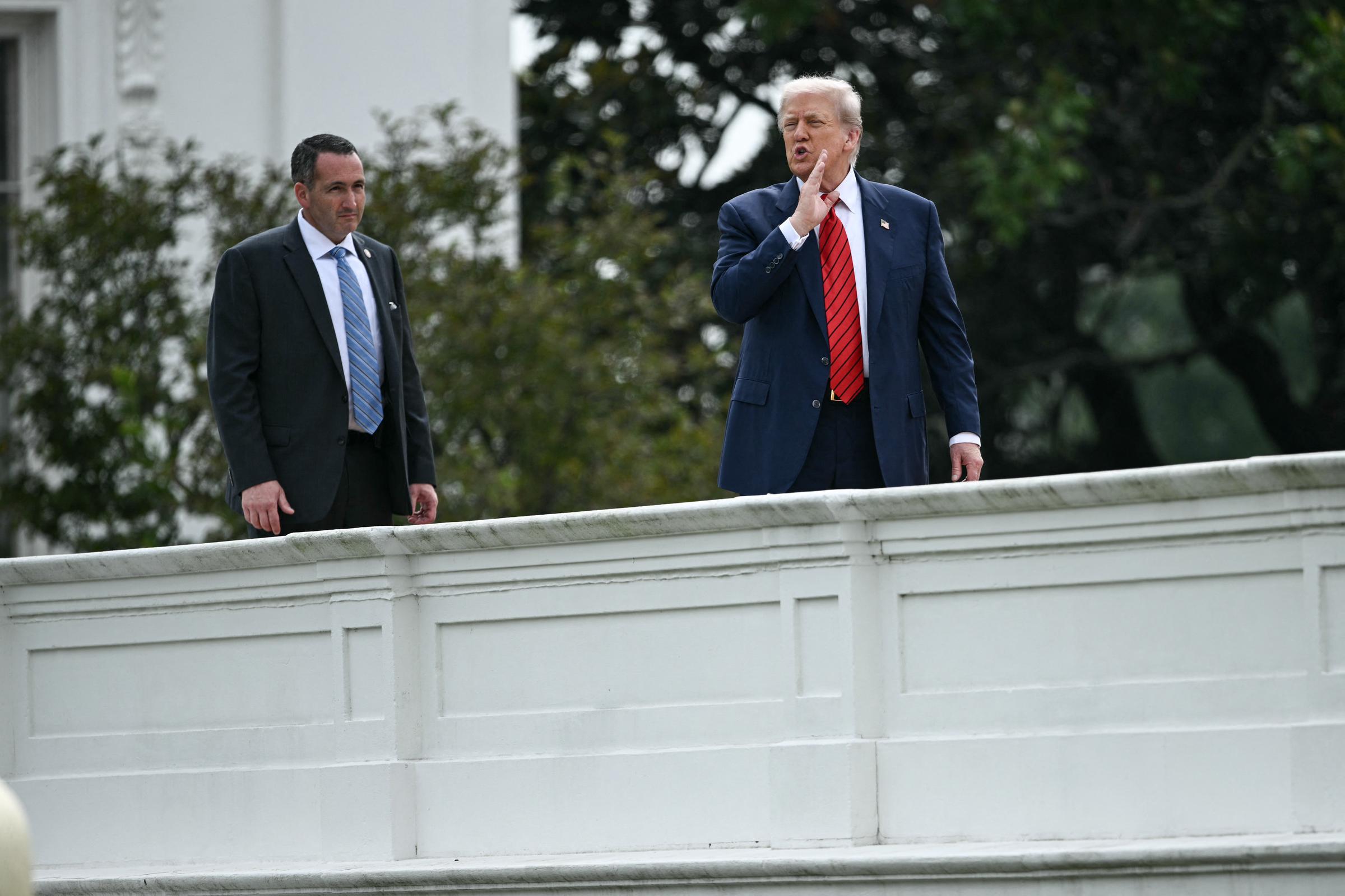 Donald Trump está de pie en el techo de la Casa Blanca, con una mano levantada hacia la boca como si llamara o se dirigiera a alguien que está abajo, con un miembro del personal de seguridad a su lado.