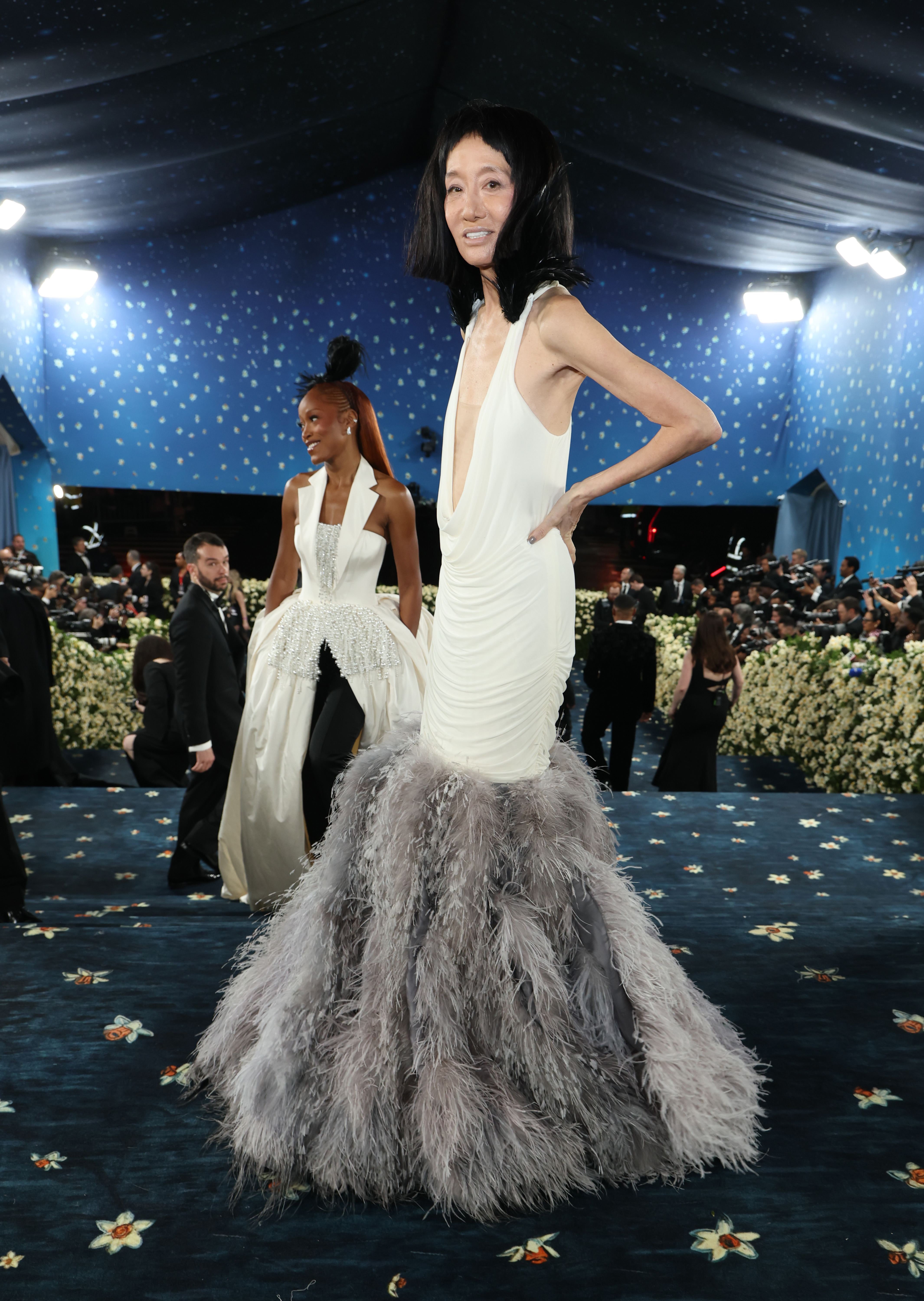 Rodeada de un mar de cámaras y decoración floral, Vera Wang posa a medio paso con su vestido texturizado mientras Keke Palmer la sigue, ambas vestidas con llamativos looks blancos con toque de plumas.