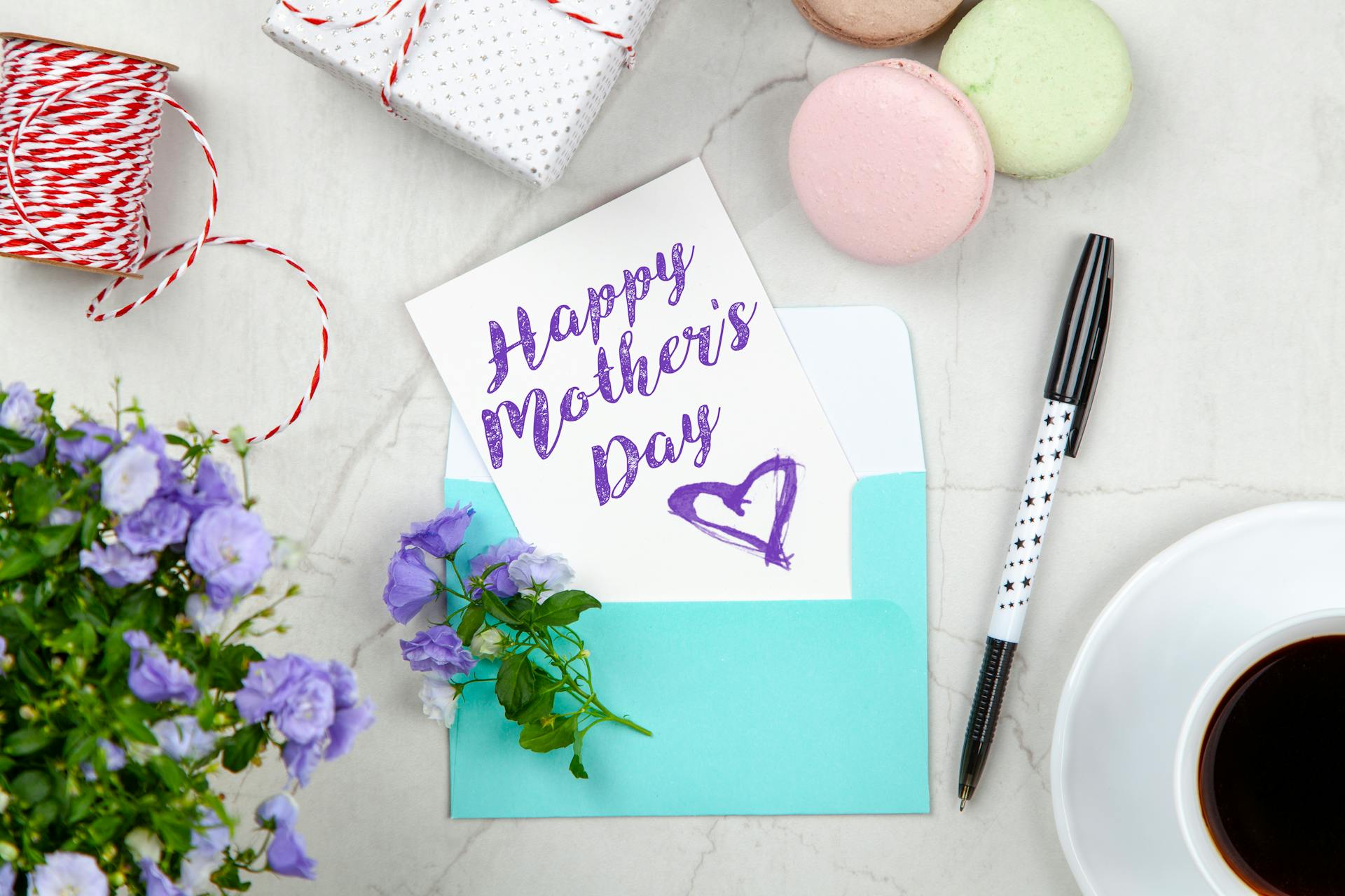 Una tarjeta del día de la madre junto a un bolígrafo, macarrones, flores y una caja cerca de una taza de café con platillo | Fuente: Pexels