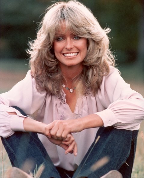 Retrato publicitario de la actriz y modelo estadounidense Farrah Fawcett sonriendo mientras está sentado al aire libre en jeans y una blusa malva. | Foto: Getty Image