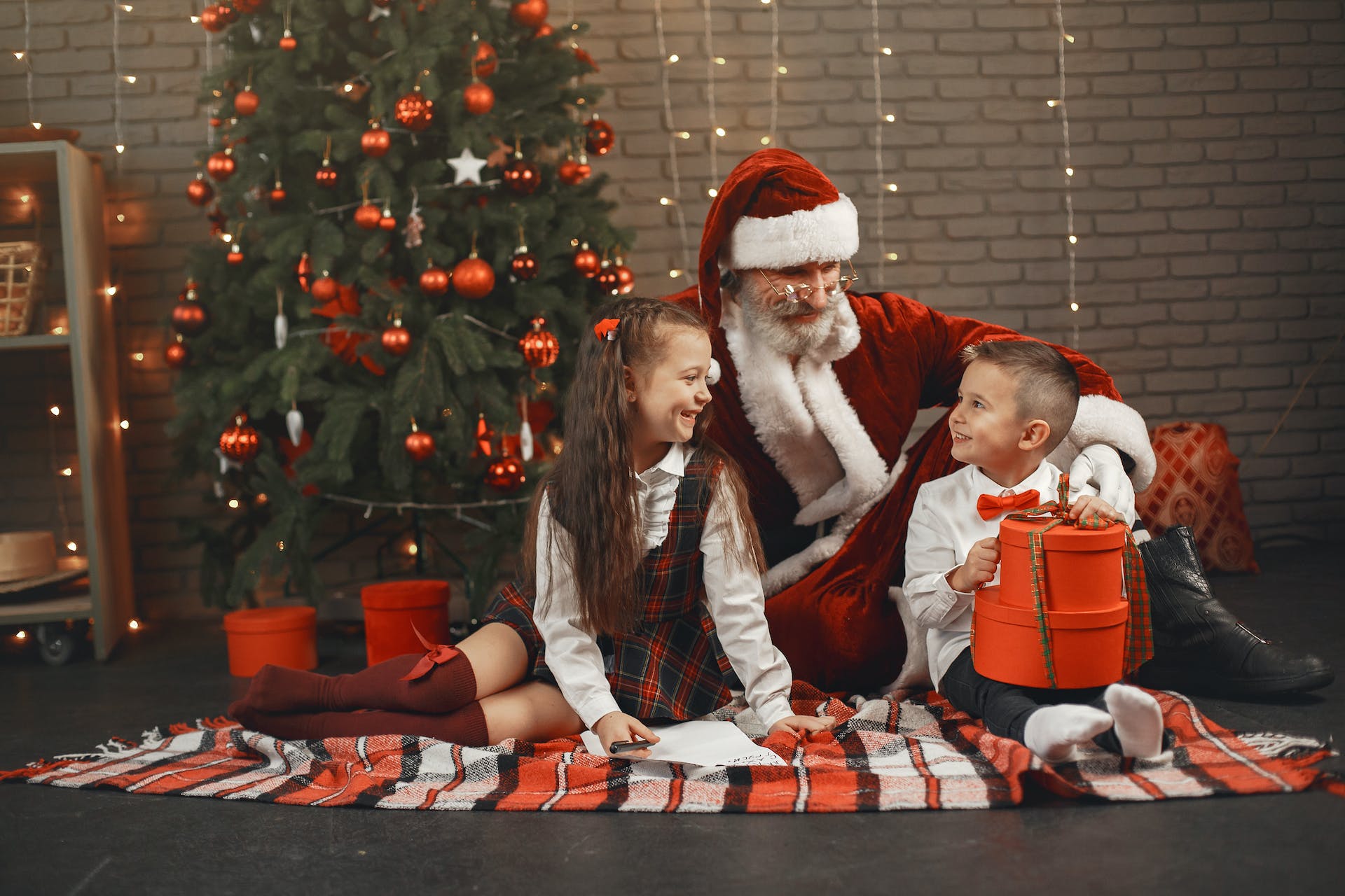 Hombre disfrazado de Papá Noel sentado con un niño y una niña junto a un árbol de Navidad | Foto: Pexels