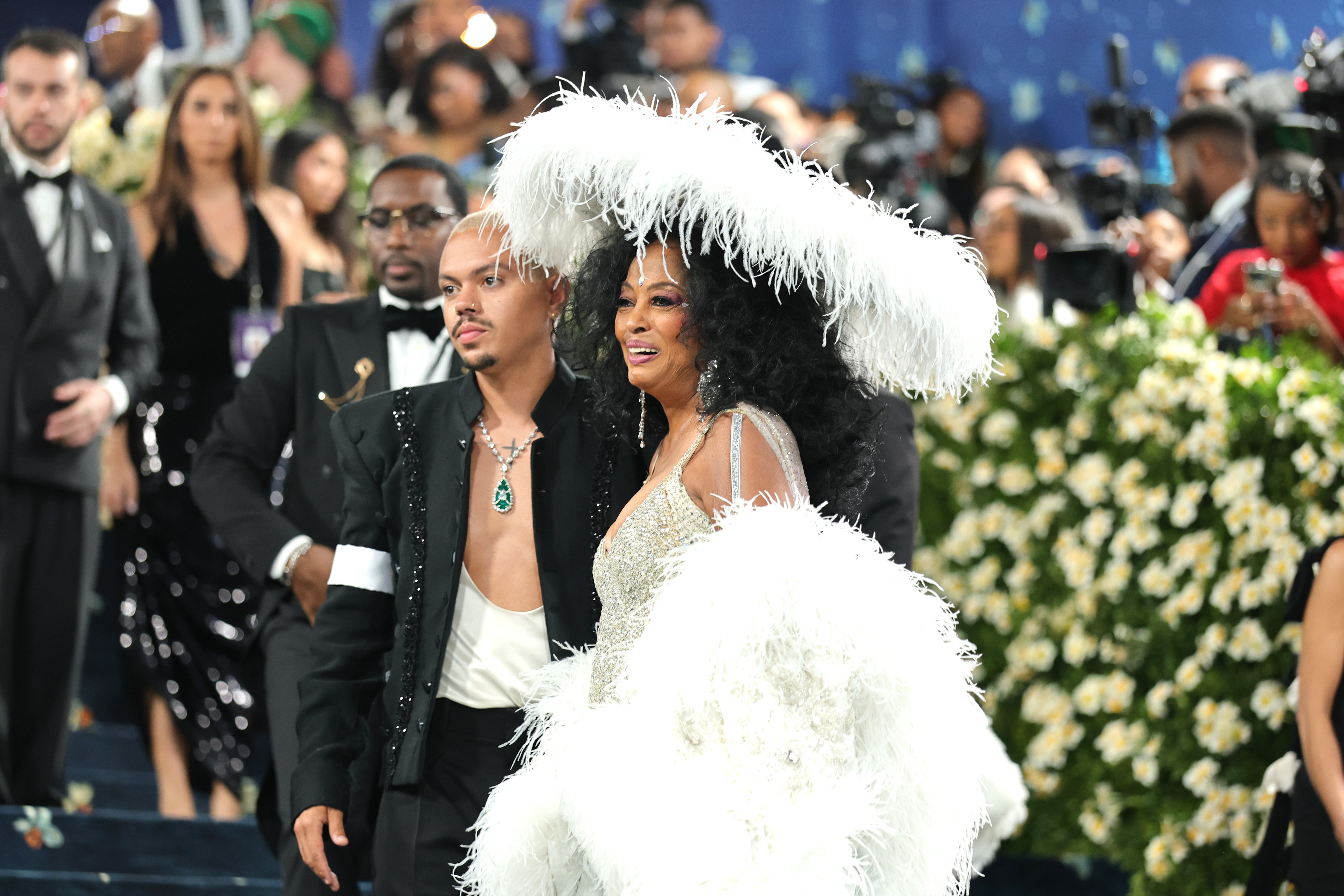 En un plano más cercano, el escote abierto de Evan Ross revela un tatuaje de una cruz en el pecho y un collar con una llamativa gota de esmeralda que descansa sobre su cuello. A su lado, su madre sonríe dulcemente, con expresión cálida y orgullosa.
