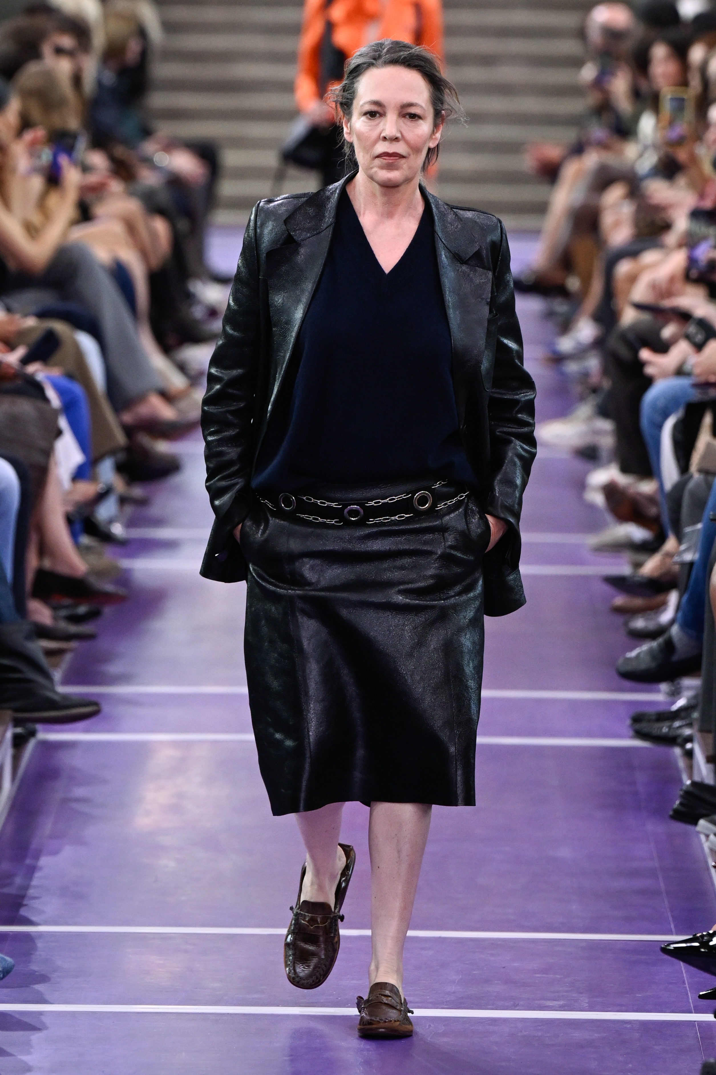 Olivia Colman desfila por la pasarela de Miu Miu en el Palais d’Iéna durante la Semana de la Moda de París, con una chaqueta de cuero negra, un top azul marino y una falda de cuero hasta la rodilla, combinada con unos mocasines marrones. | Fuente: Getty Images