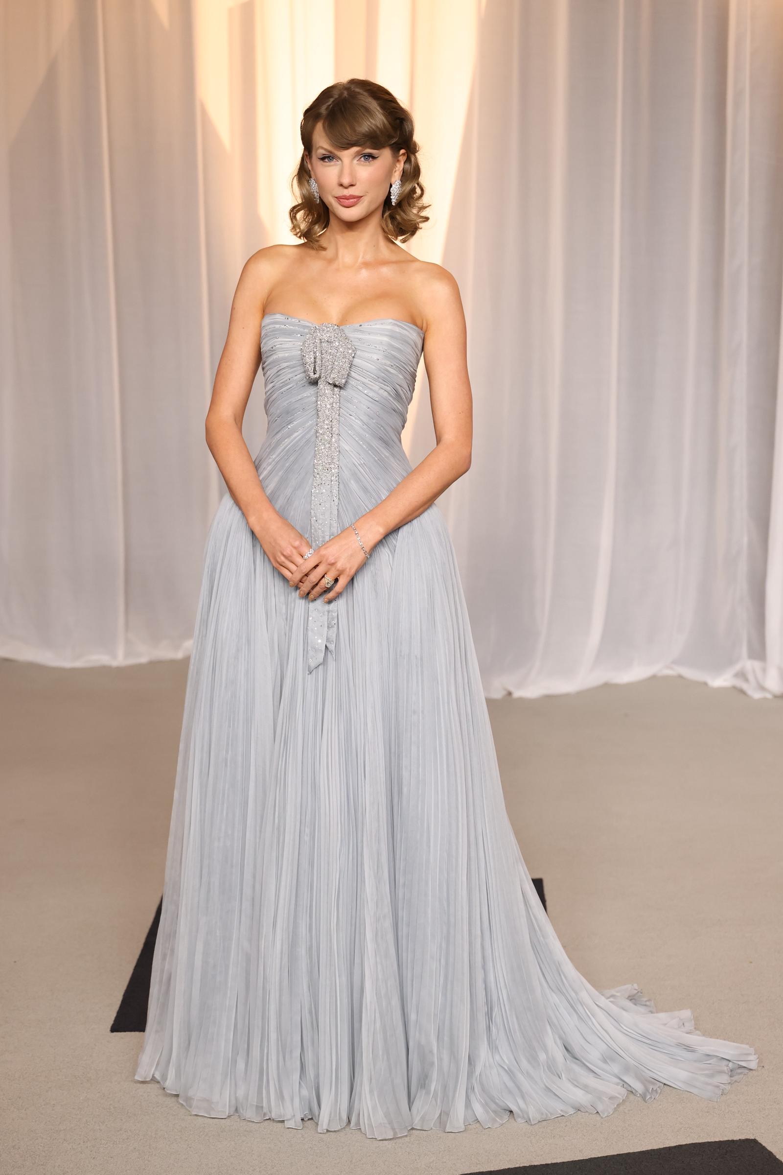 Taylor Swift posa con un vestido sin tirantes, plisado y de color azul hielo, con un lazo cubierto de cristales y una larga cola, y lo complementa con unos pendientes brillantes, una pulsera y unos anillos.