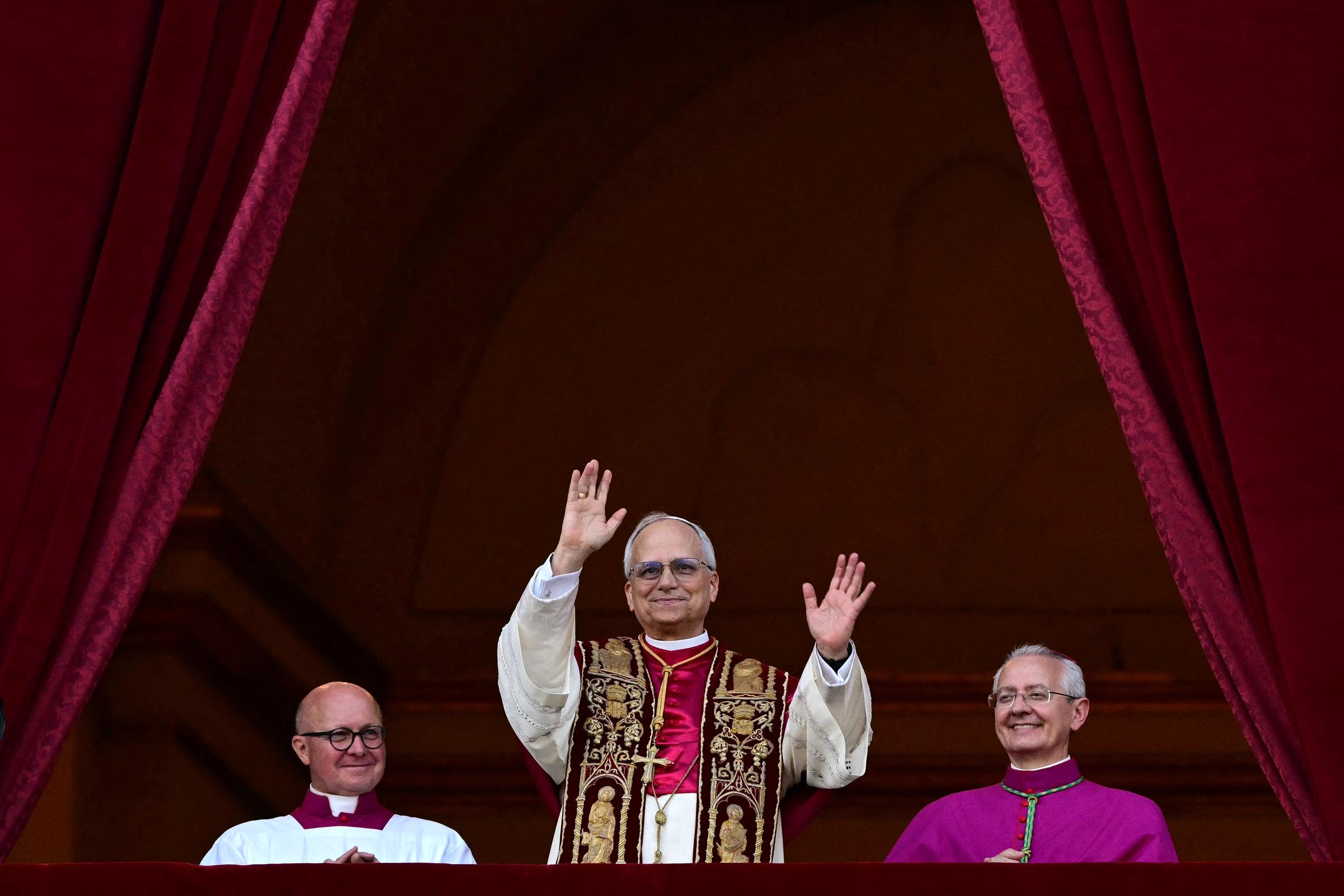 El papa León XIV levanta ambas manos en señal de saludo, lleva una estola ricamente bordada sobre una túnica color crema y sale al balcón enmarcado por cortinas de terciopelo rojo intenso.