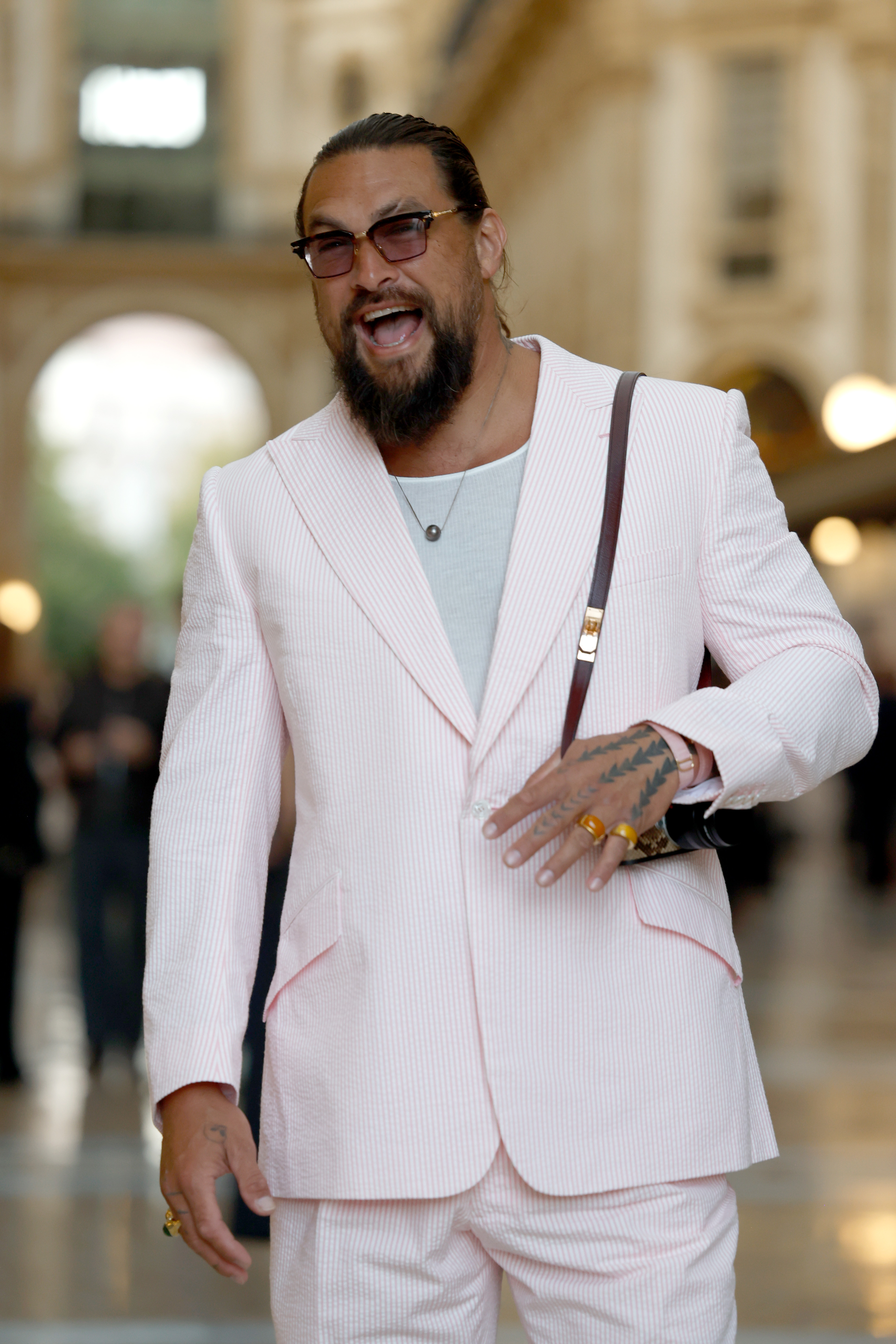 Momoa se ríe mientras posa dentro del recinto, lo que nos permite ver más de cerca sus gafas tintadas, sus joyas y las finas rayas rosas que recorren su traje. | Fuente: Getty Images