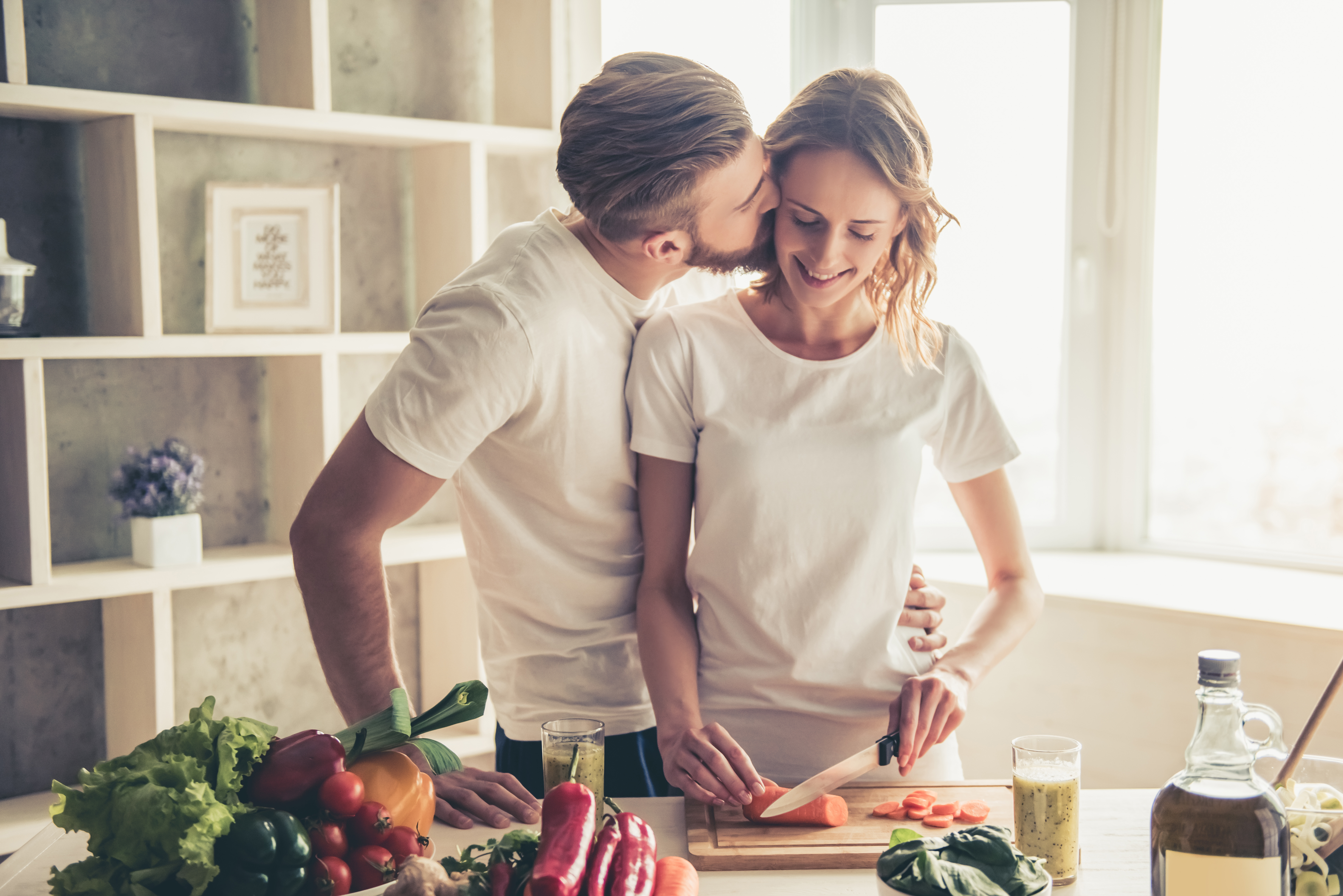 Un hombre besa a su mujer mientras ella corta verduras | Fuente: Shutterstock